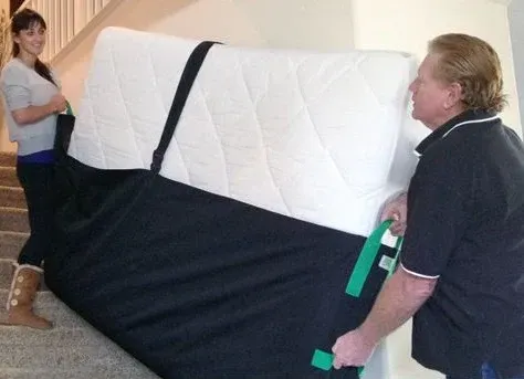 Two people carrying a mattress up stairs with black lifting straps.