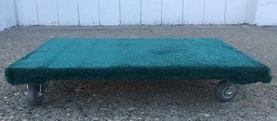 A rectangular, green-carpeted cart with four wheels sits on a gravel surface.