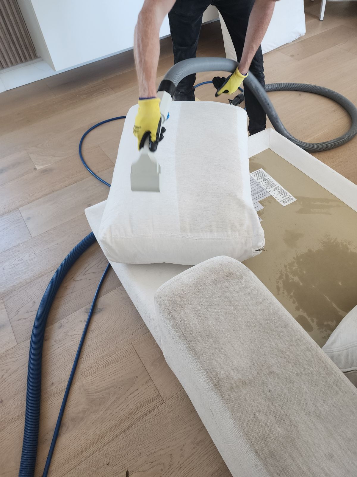 Person cleaning a white upholstered chair with a carpet cleaner in a room with a rug and hose.