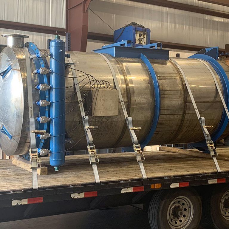 Large stainless steel industrial tank secured with blue straps on a flatbed trailer inside a warehouse.