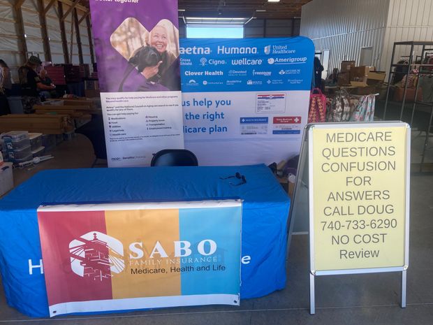 A booth for Sabo Insurance with Medicare information, a blue table cover, and a sign to call Doug for no-cost help.