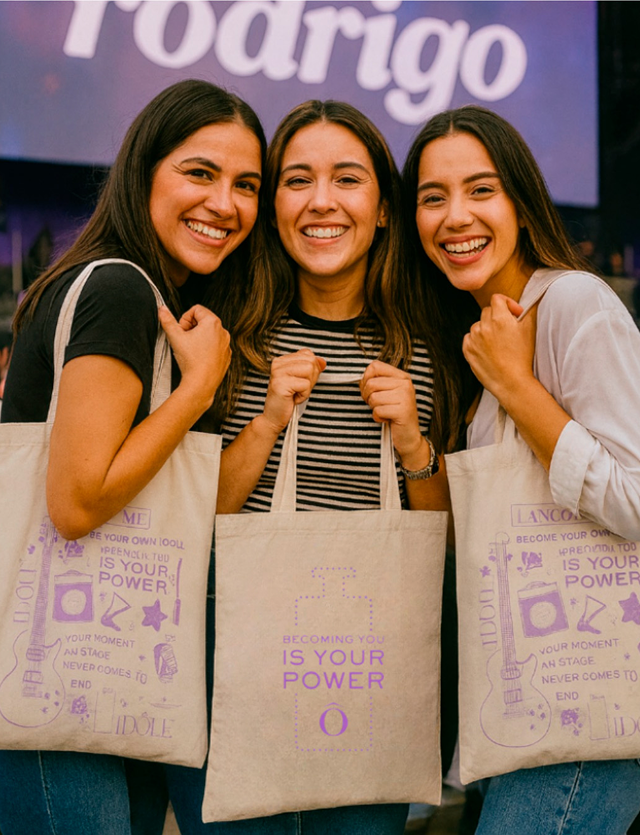 Tres mujeres sonrientes posan juntas sosteniendo bolsas de tela en un stand de un evento.