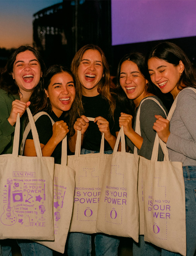 Cinco personas sonrientes posan en un evento, sosteniendo bolsas de tela color beige con texto morado.