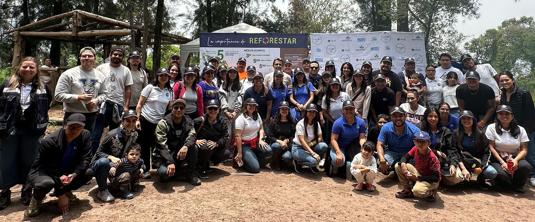 Un grupo numeroso posa al aire libre frente a pancartas y árboles; algunos están sentados y otros de pie.