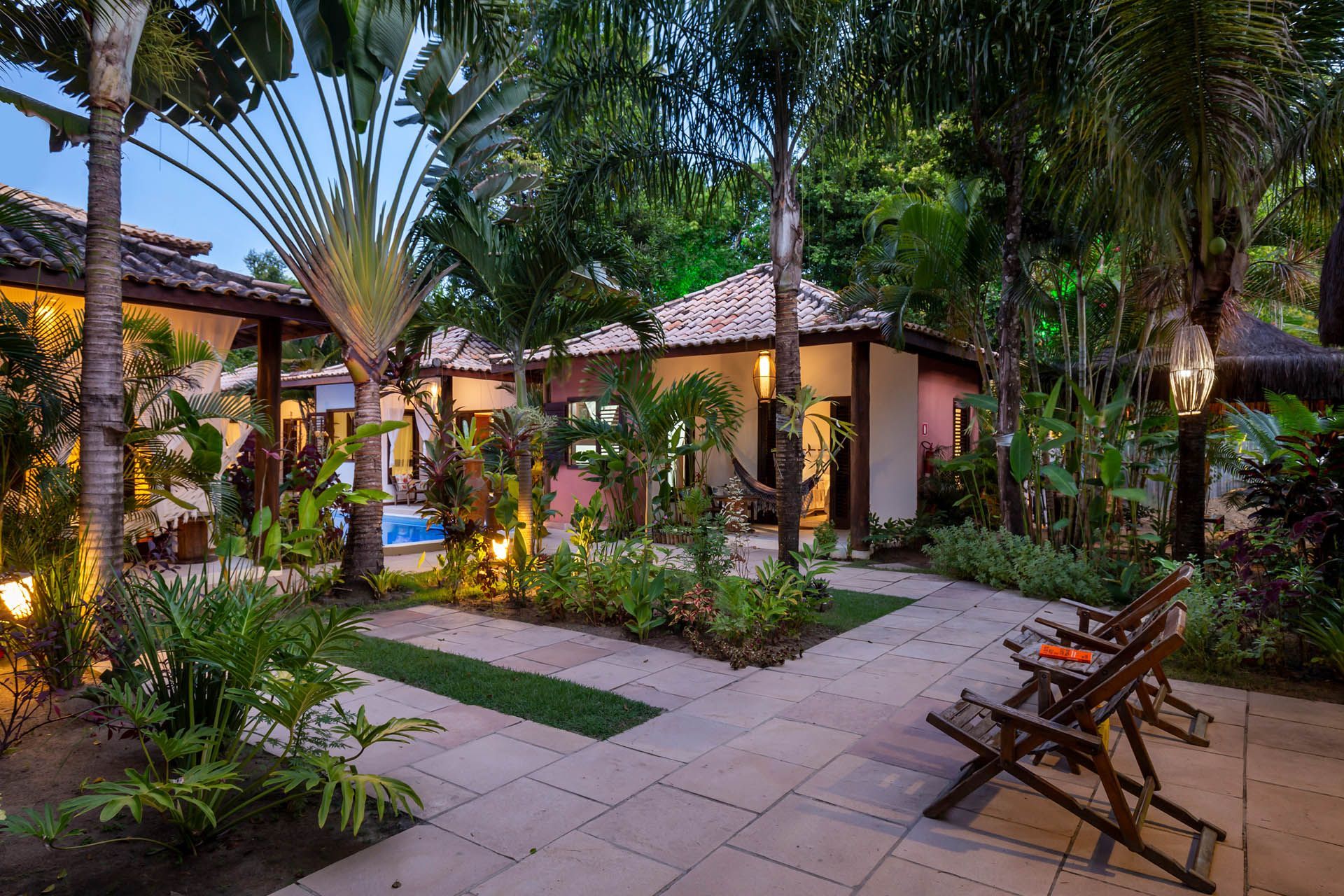 Jardim tropical com pátio, palmeiras, caminhos de azulejo e espreguiçadeiras ao entardecer.