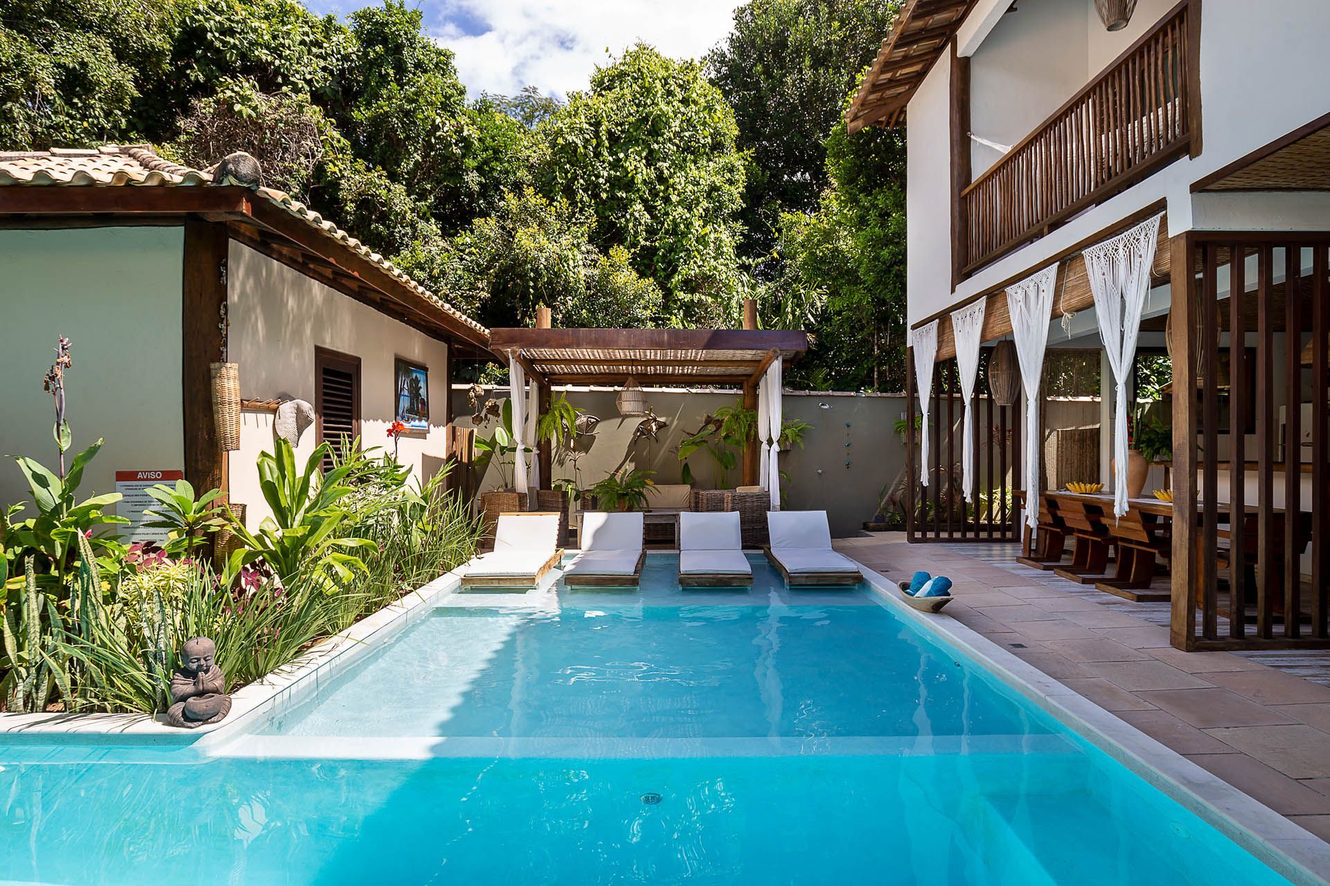 Piscina de resort tropical com espreguiçadeiras, cabana e vegetação exuberante ao lado de uma villa.