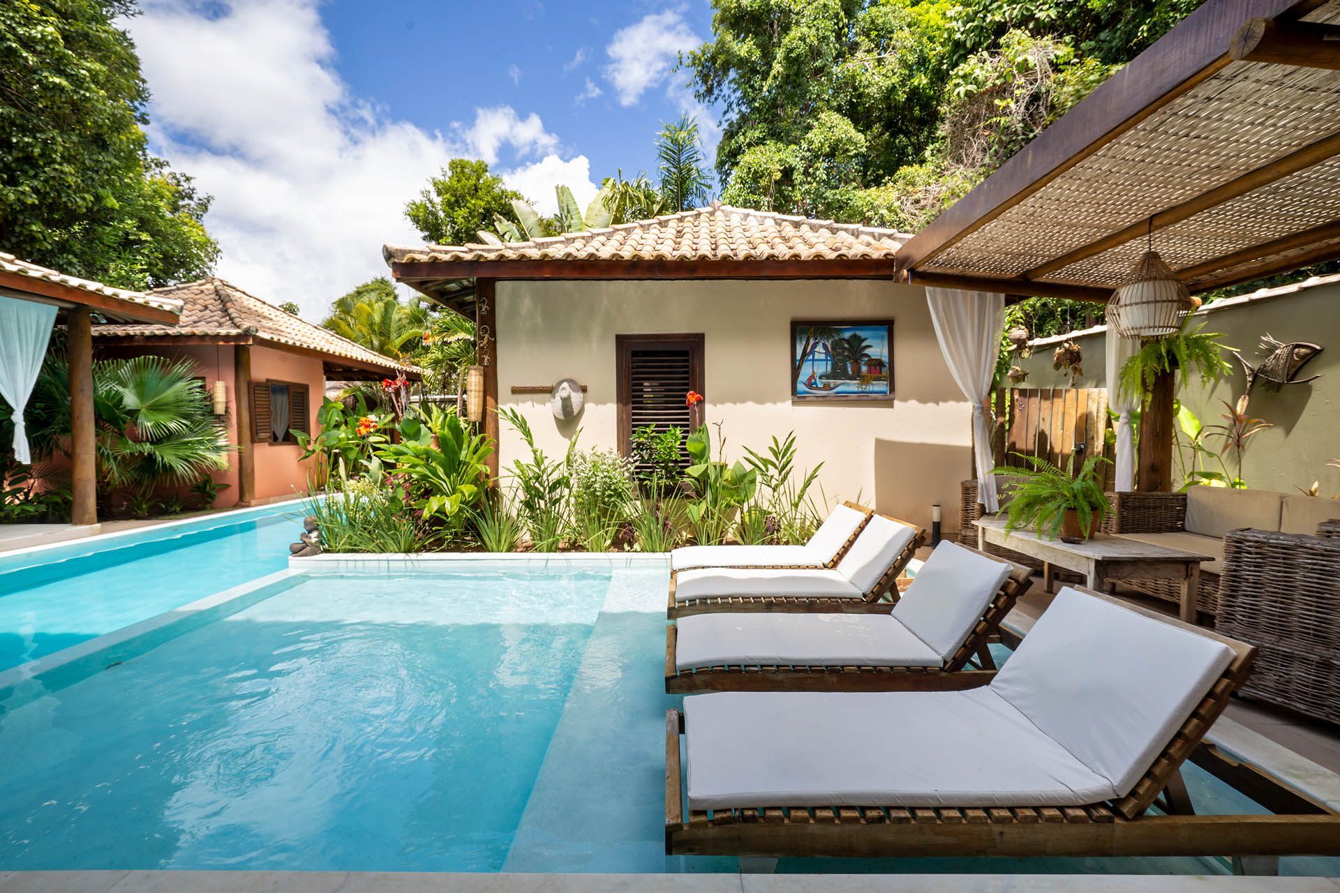 Vila tropical à beira da piscina com espreguiçadeiras brancas, guarda-sóis de palha e um céu azul ensolarado.