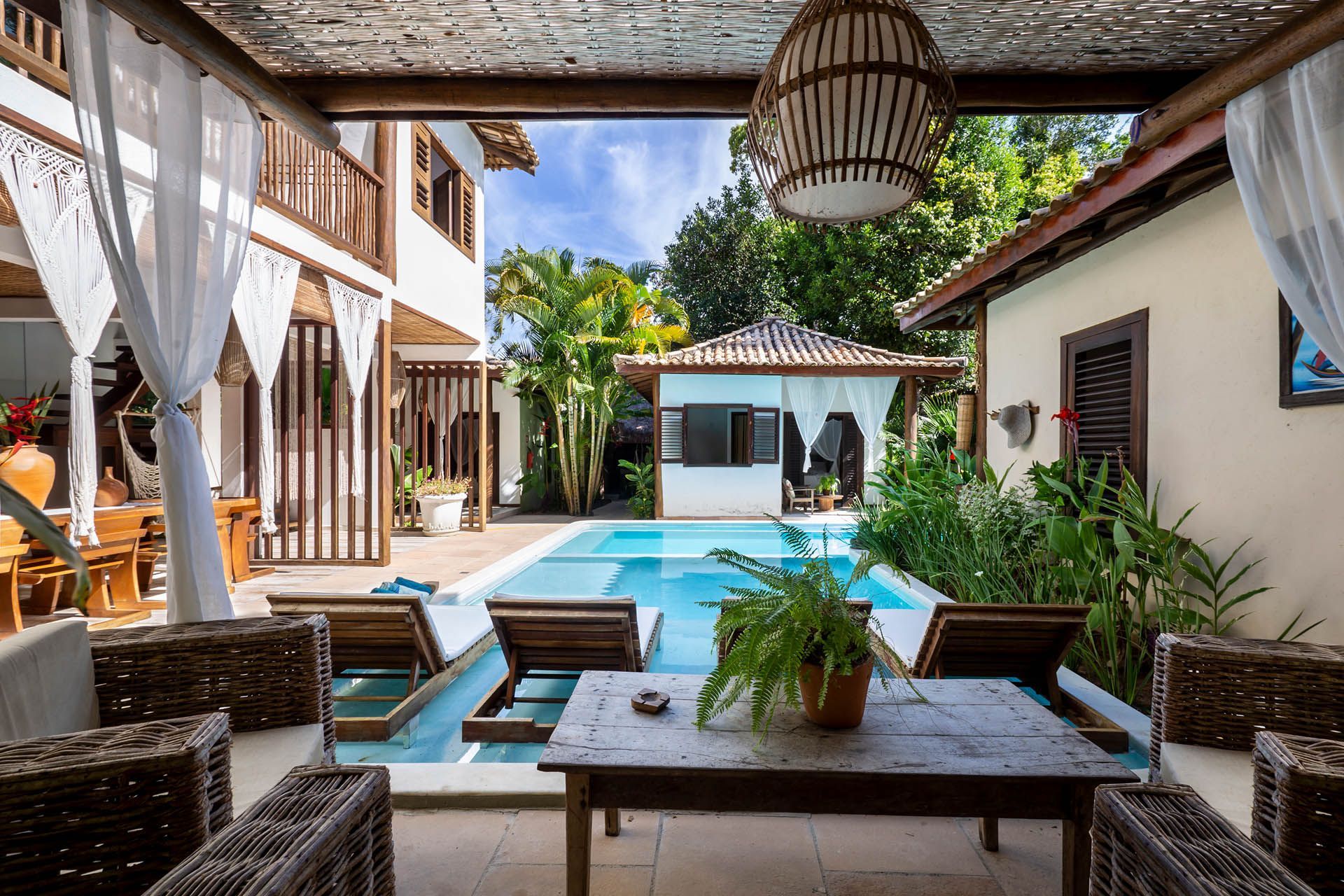 Pátio coberto com vista para uma piscina azul e uma casa branca, com cadeiras de vime e plantas tropicais exuberantes.