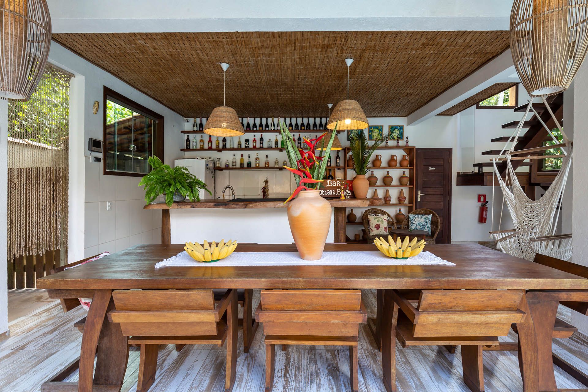 Sala de jantar rústica com mesa de madeira, cadeiras de vime, luminárias pendentes e prateleiras abertas com peças de cerâmica.