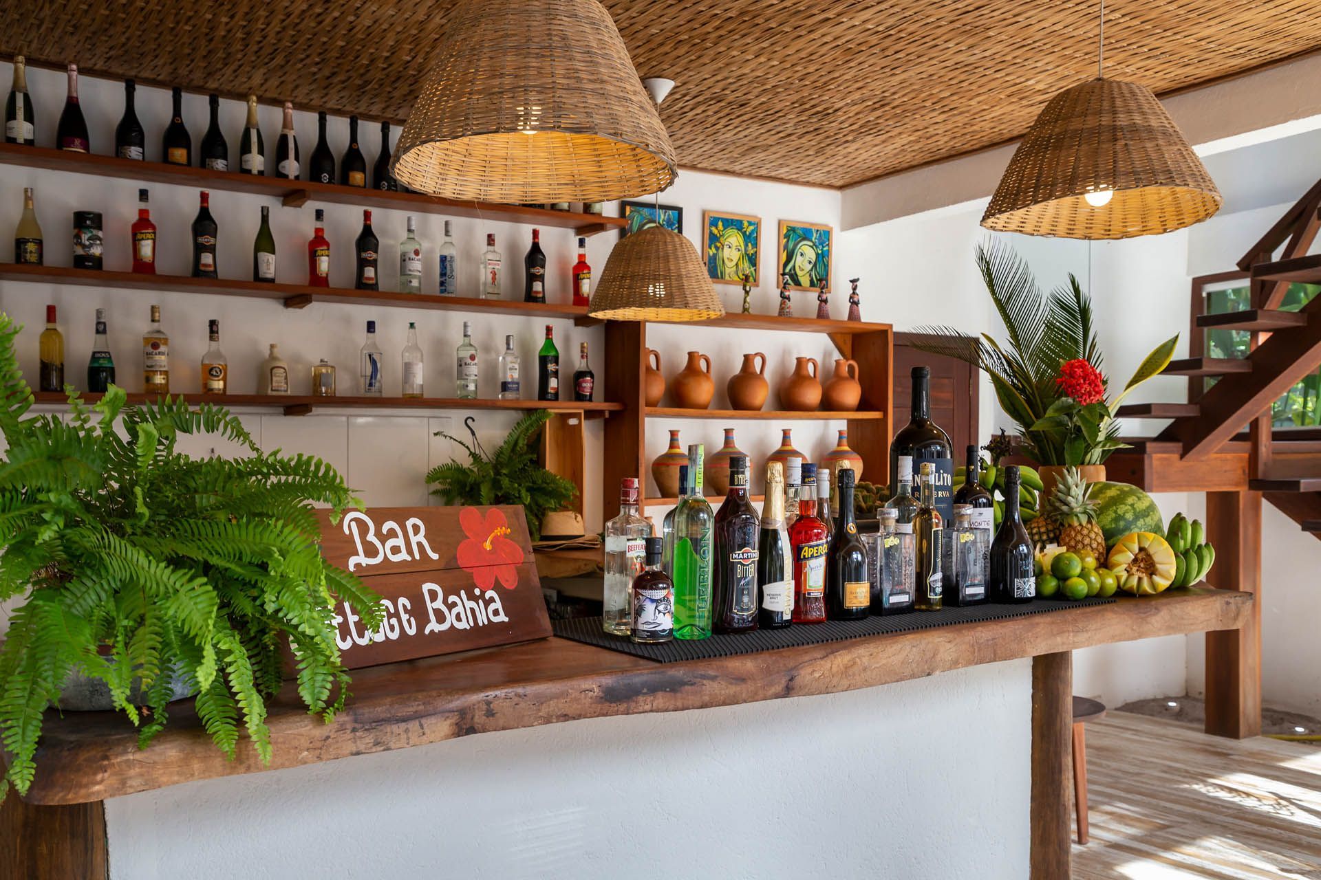 Balcão de bar rústico com garrafas de bebidas, luminárias pendentes, plantas exuberantes e uma pequena placa de lousa em um ambiente iluminado.