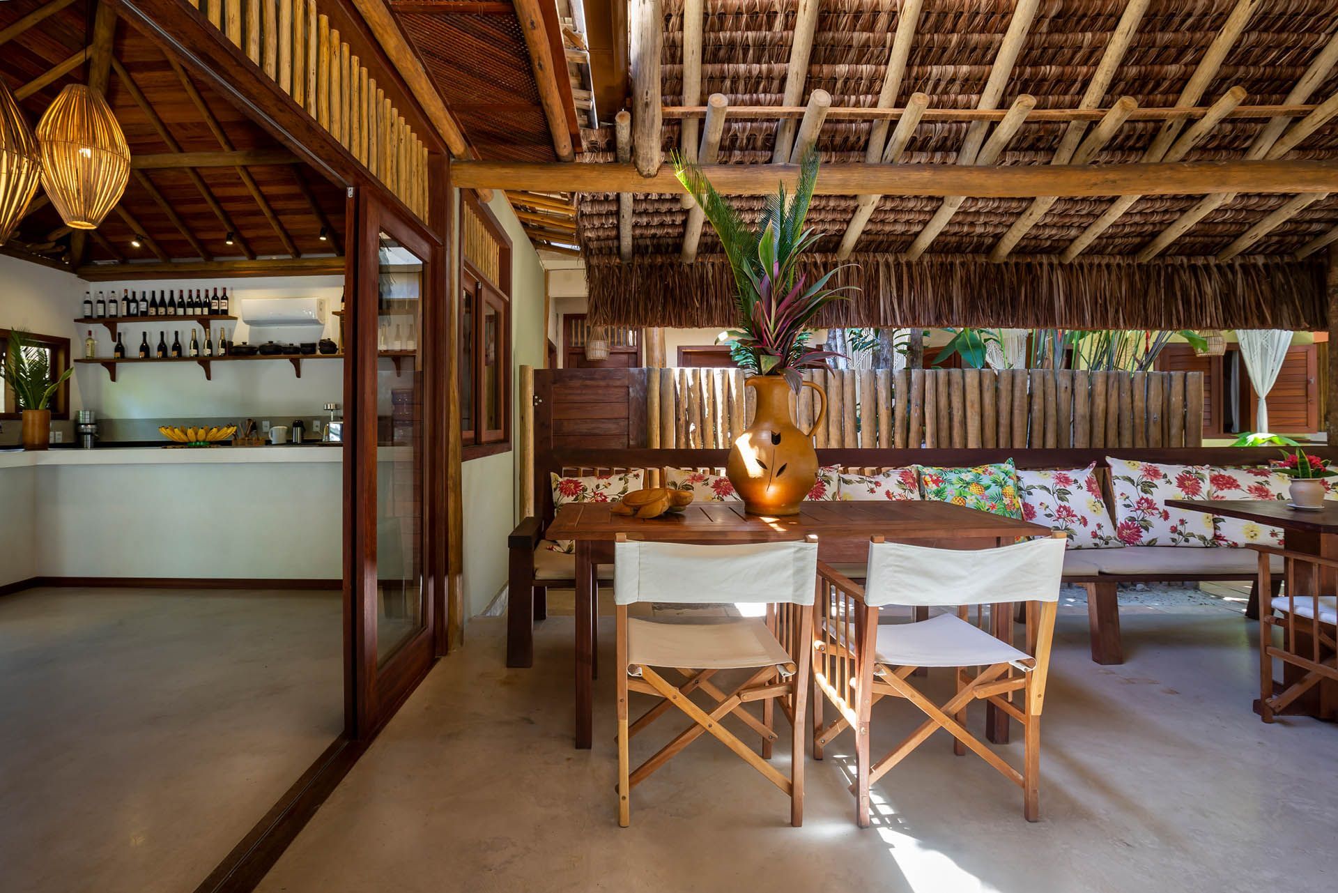 Sala de jantar tropical com mesa e cadeiras de madeira e teto de palha em um espaço amplo e iluminado.
