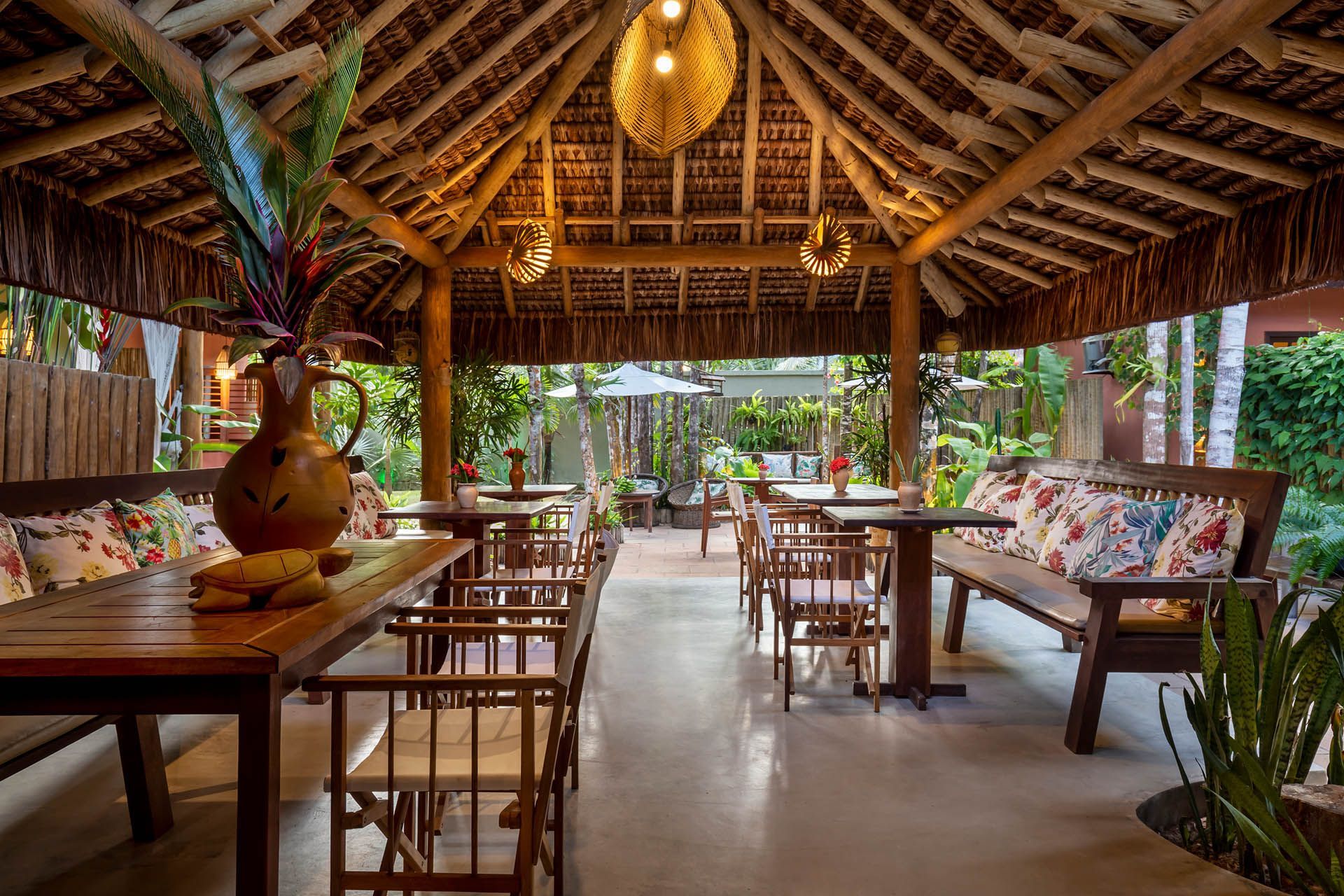 Aconchegante lounge tropical ao ar livre com mesas de madeira, bancos almofadados e teto de palha sob luzes pendentes que aquecem o ambiente.