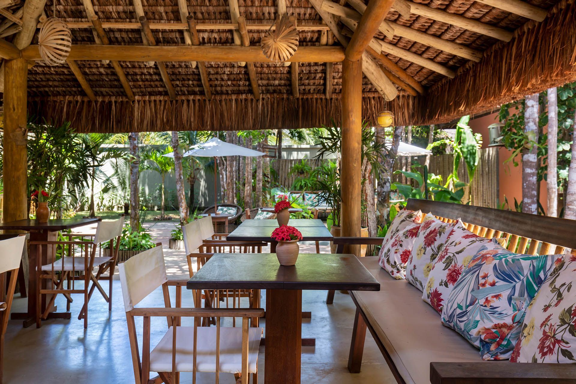 Café tropical ao ar livre com teto de palha, mesas de madeira e bancos almofadados com motivos florais.