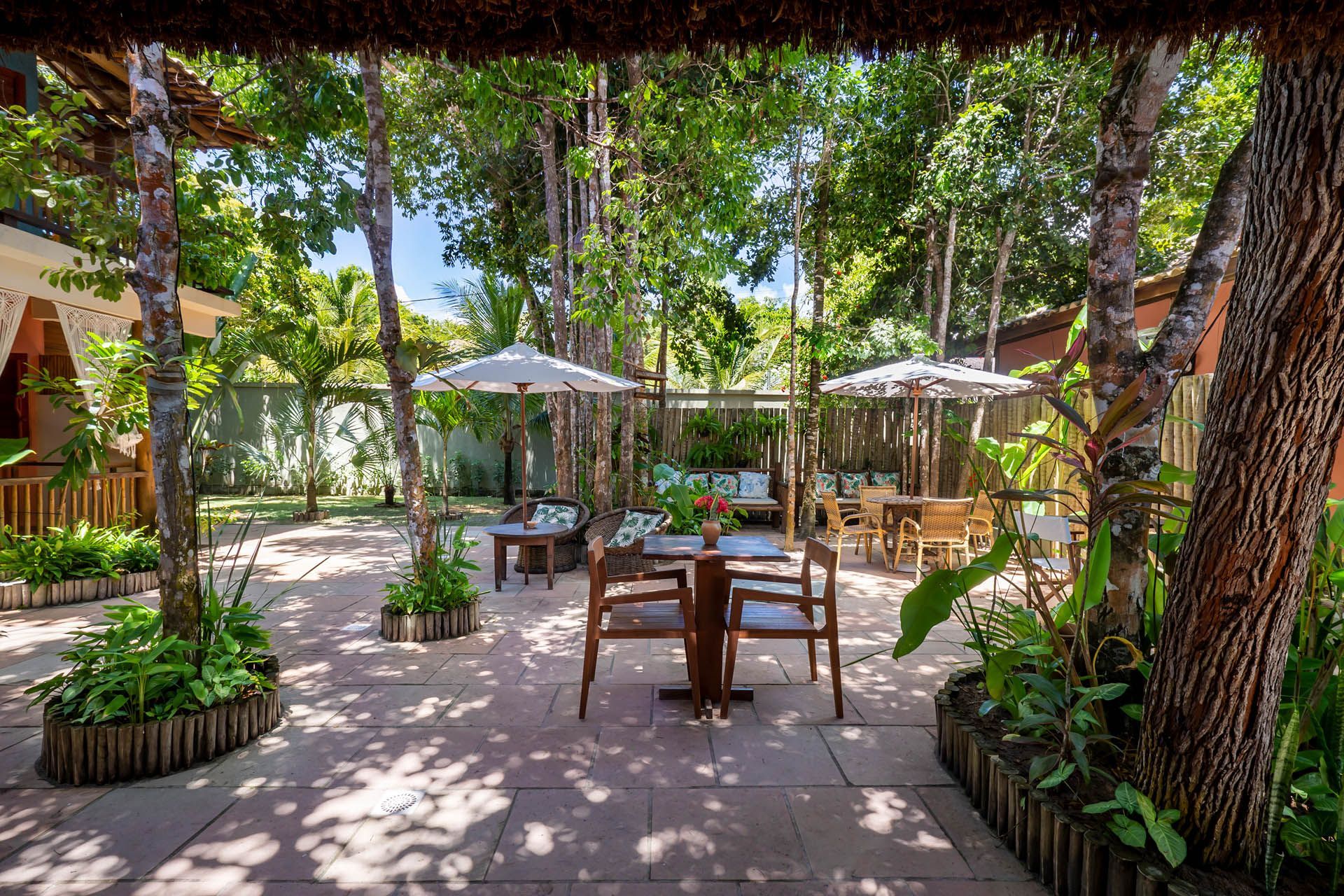 Pátio sombreado do café no jardim, com mesas, cadeiras, guarda-sóis e vegetação exuberante.