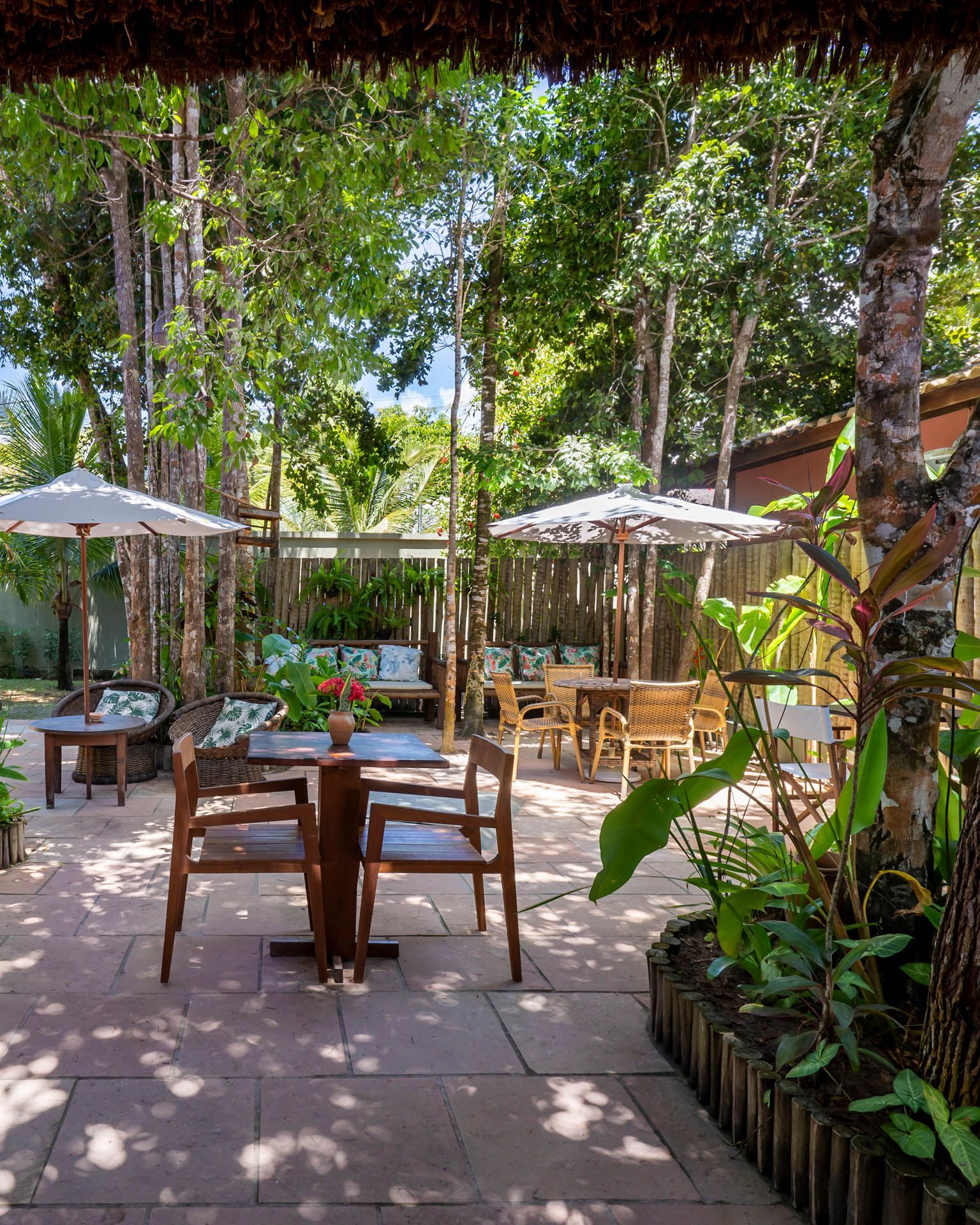 Pátio externo sombreado de um café com mesas de madeira, guarda-sóis brancos e uma vegetação exuberante, composta por árvores e plantas.