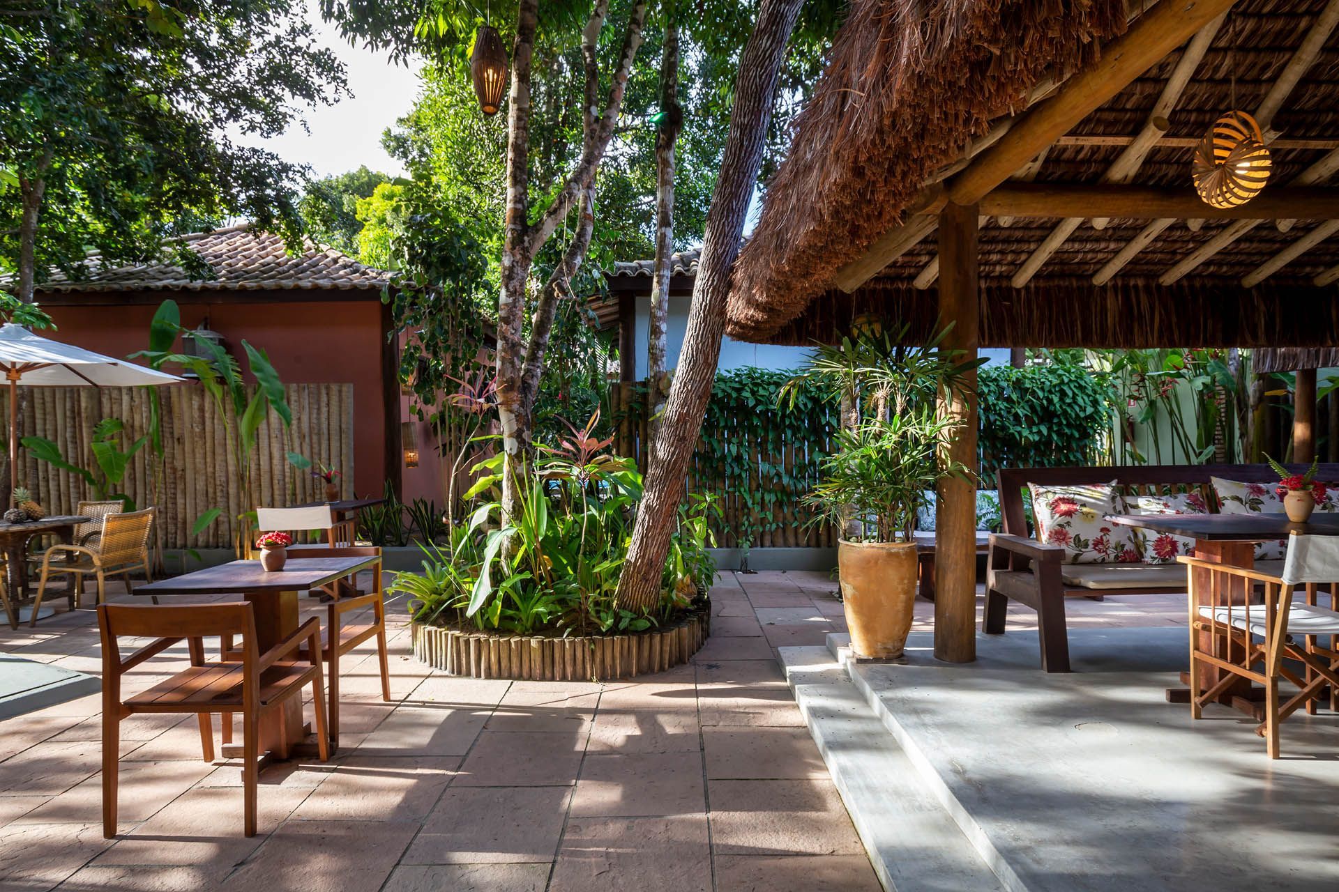 Pátio externo de café tropical com mesas e cadeiras de madeira, e vegetação sob cobertura de palha.
