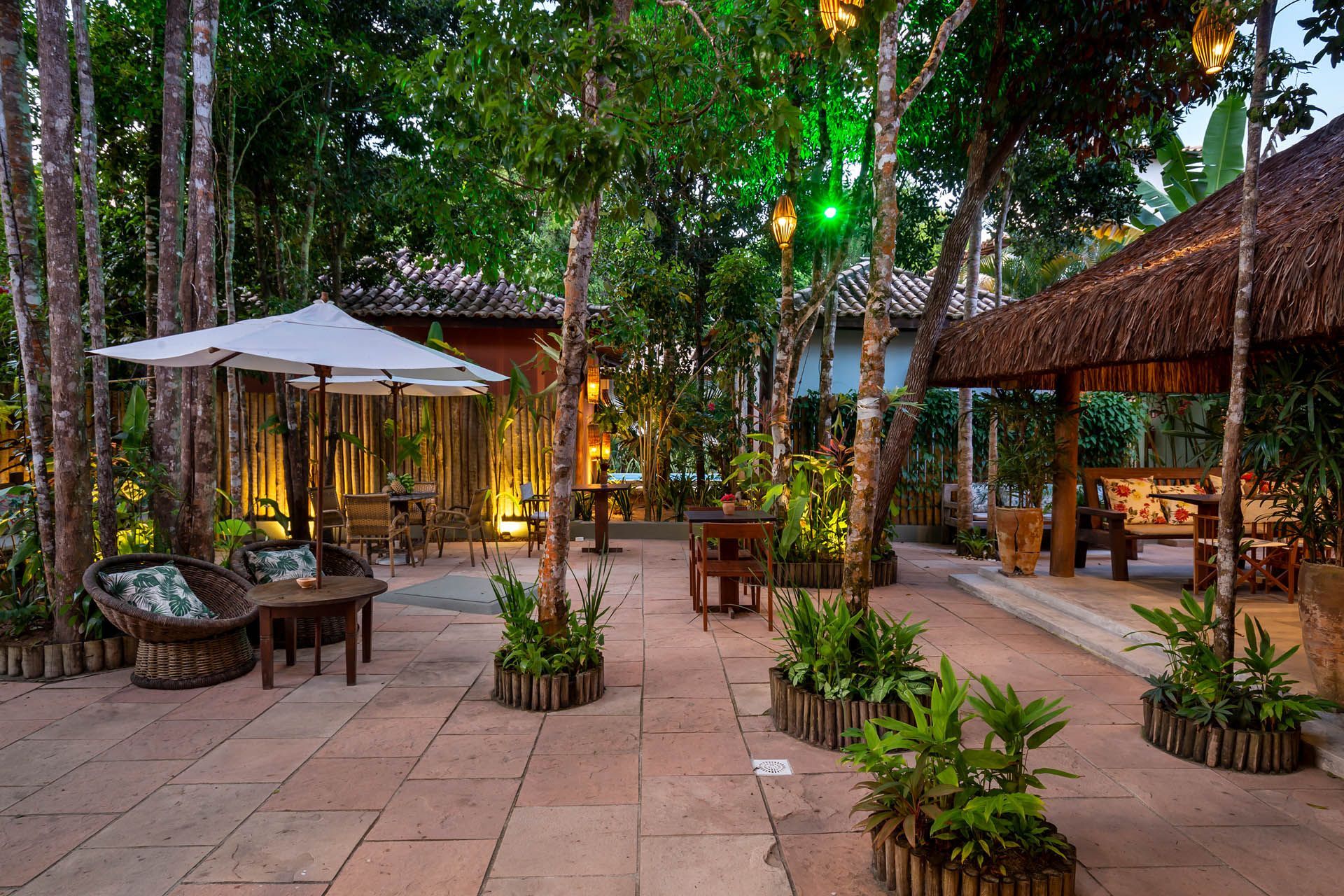 Pátio externo com mesas, cadeiras, vasos de plantas e luzes aconchegantes sob árvores altas ao entardecer.