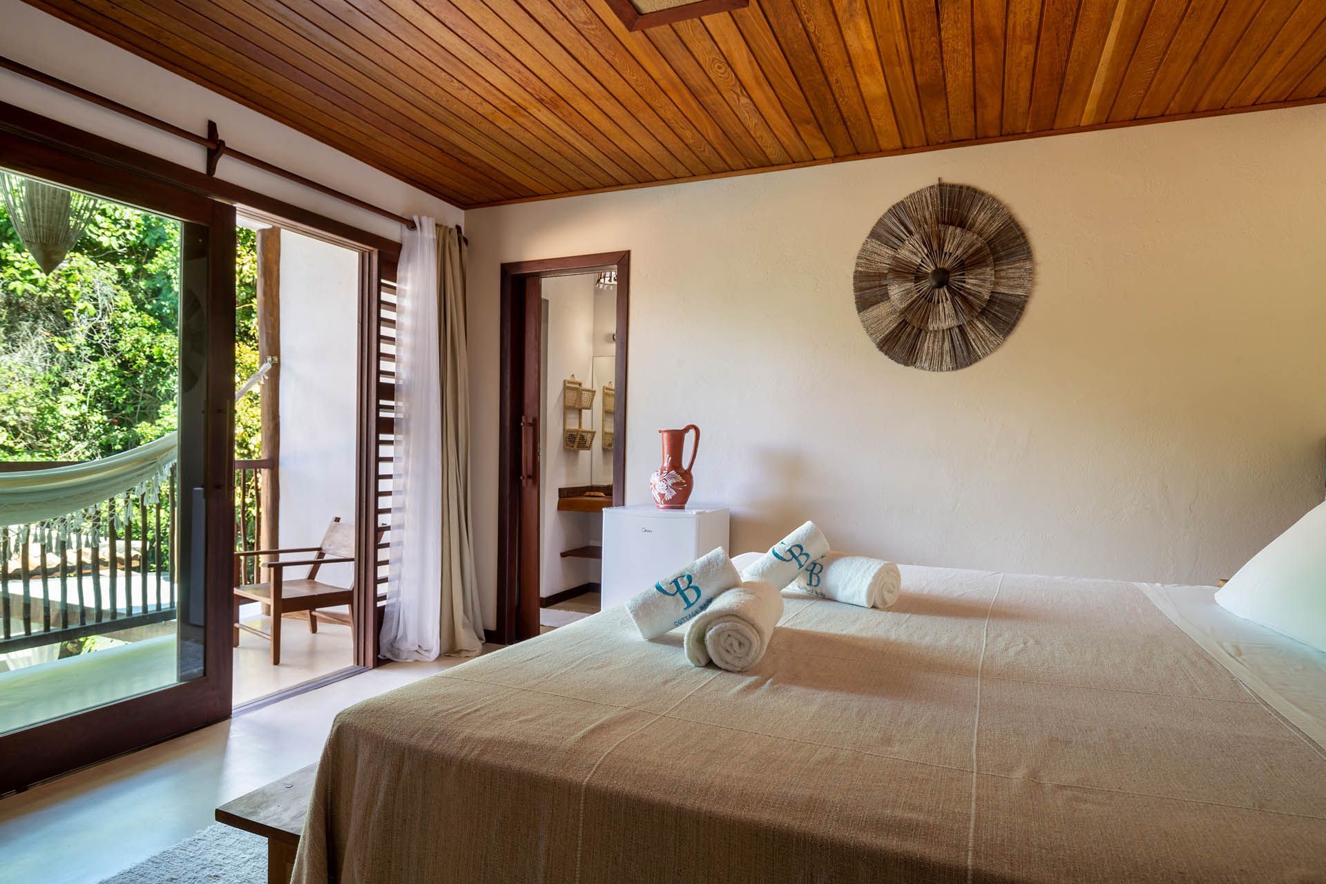 Quarto luminoso com cama, portas de varanda abertas, teto de madeira e vista para o mar ou para o jardim.