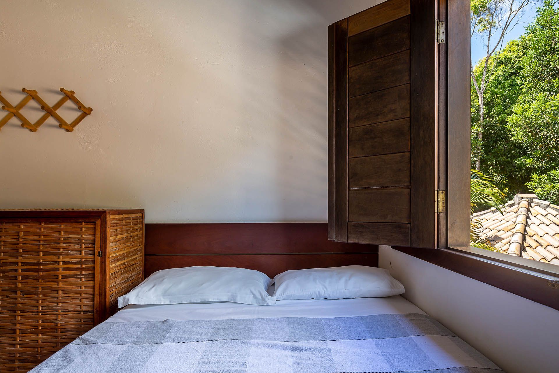 Quarto arrumado com cama branca, cômoda de vime e janela aberta com vista para a vegetação.