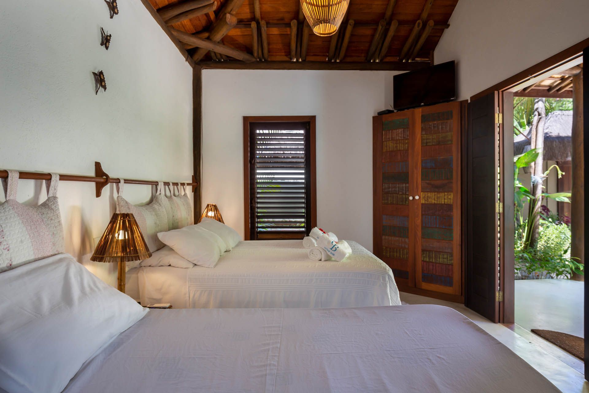 Quarto luminoso com duas camas, teto de madeira e porta aberta para um jardim tropical.