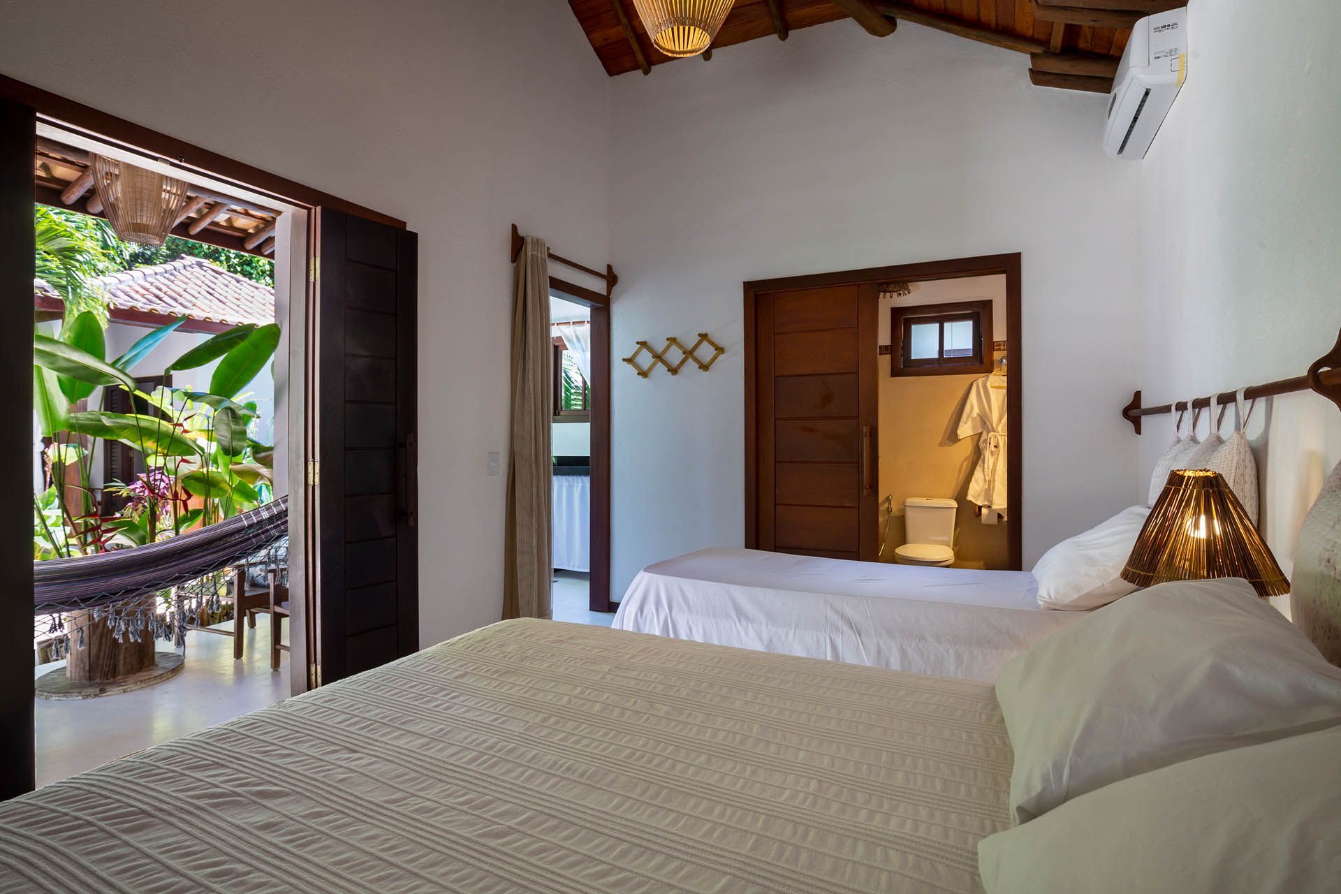 Quarto luminoso com cama branca, teto de madeira, porta de varanda aberta e vegetação tropical no exterior.