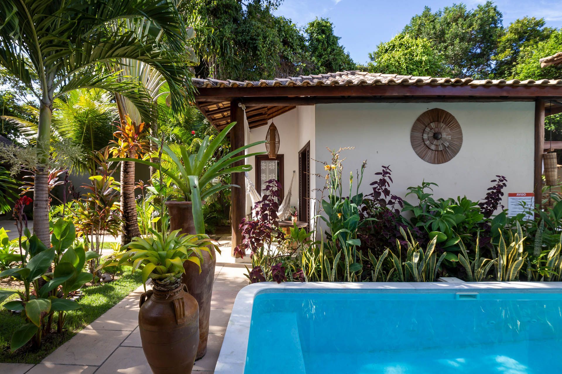 Pátio tropical com piscina azul, plantas exuberantes e uma casa branca sob um telhado de telhas.