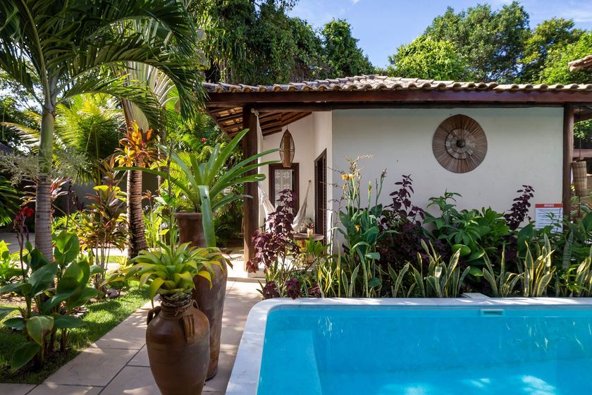 Pátio tropical com piscina azul, plantas exuberantes e uma casa branca sob um telhado de telhas.