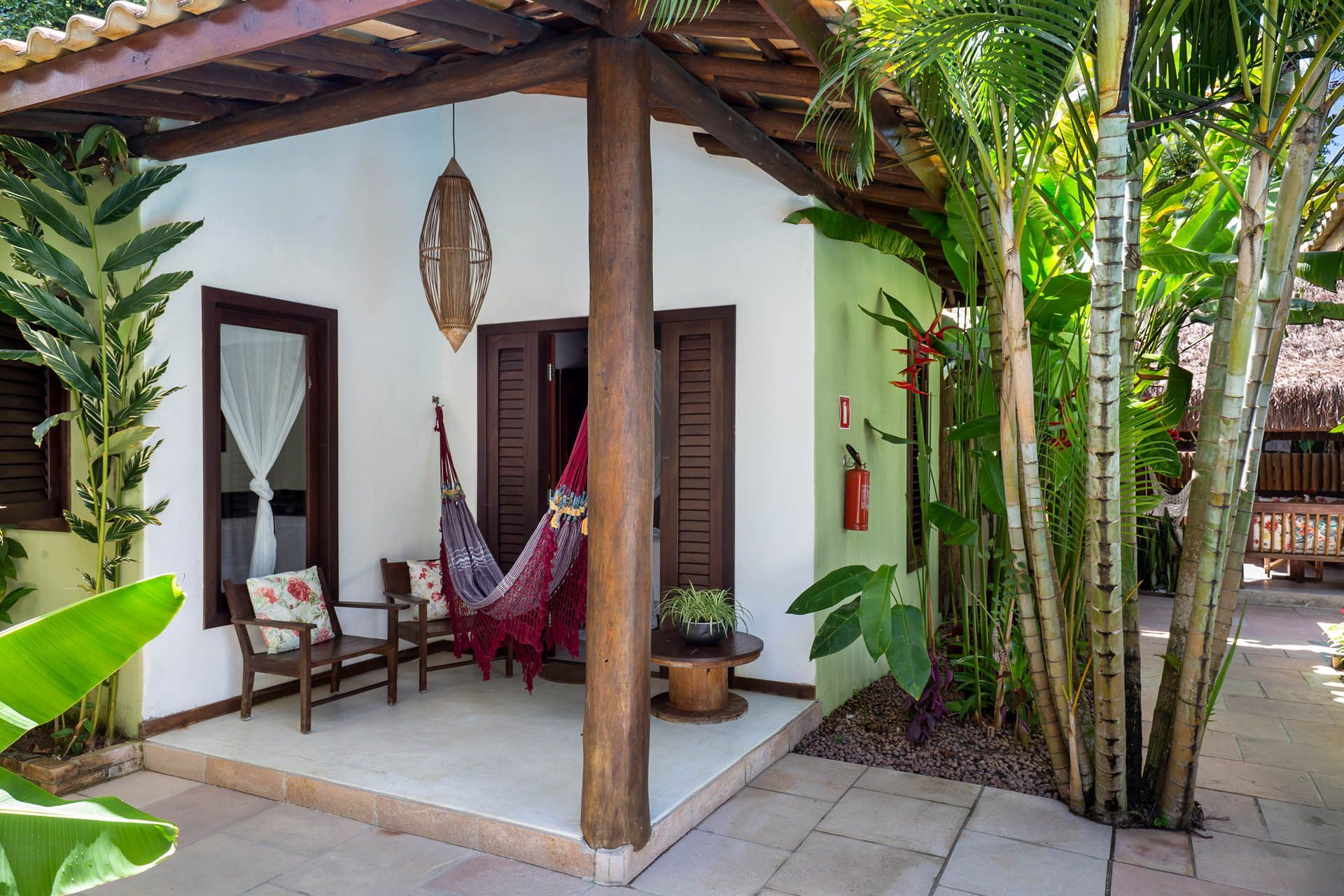 Varanda tropical com rede, cadeiras de madeira e vegetação exuberante ao lado de uma casa branca.