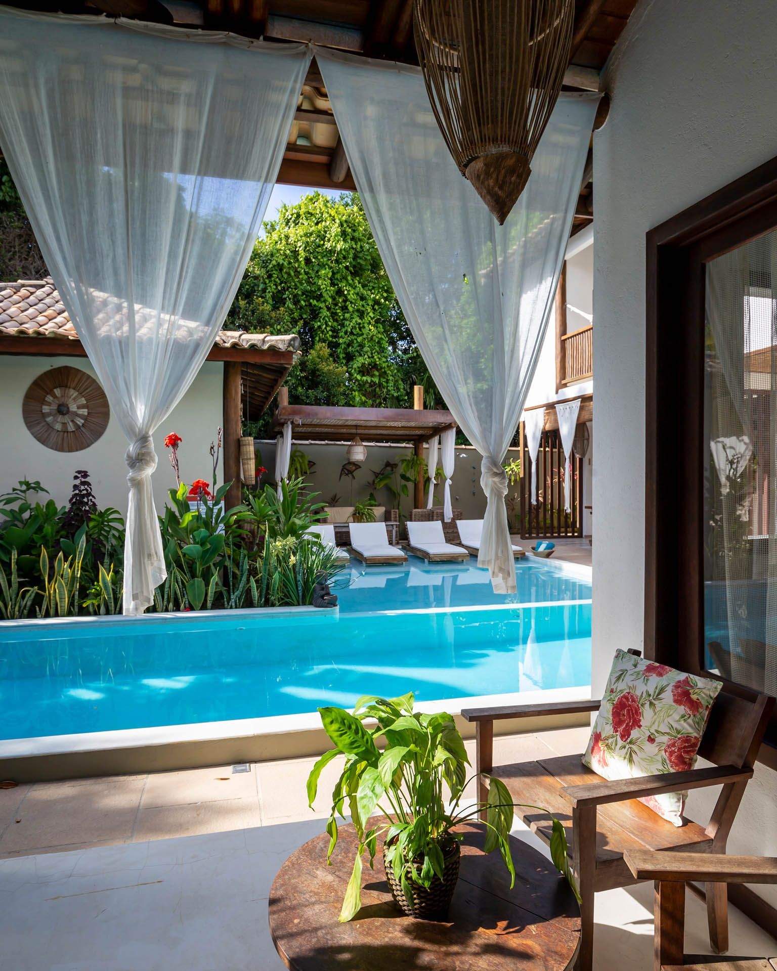 Pátio coberto com vista para uma piscina turquesa, com espreguiçadeiras e plantas tropicais, emoldurado por cortinas brancas.