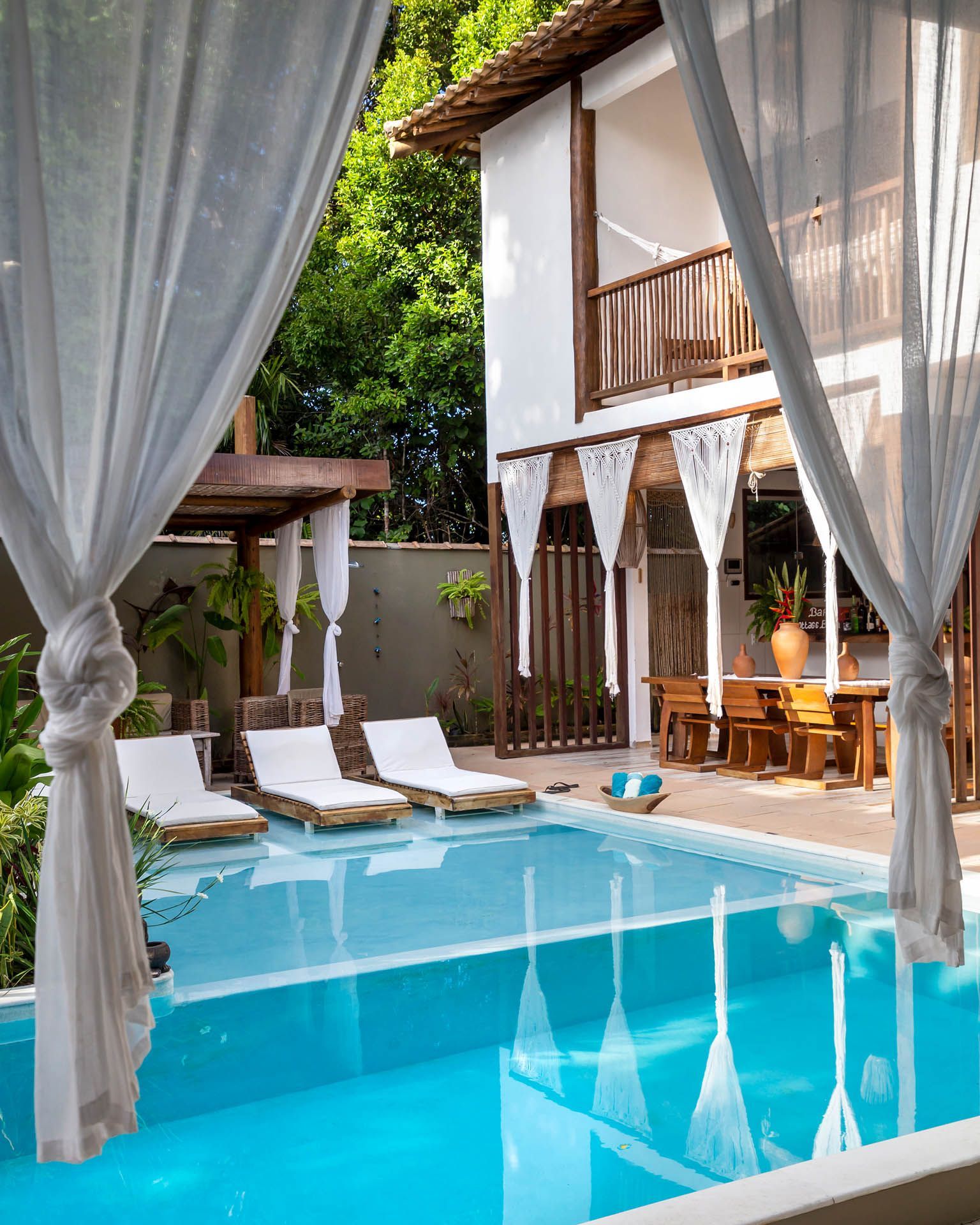 Pátio de piscina luxuoso com espreguiçadeiras brancas, cortinas drapeadas e varanda em um cenário de resort tropical.
