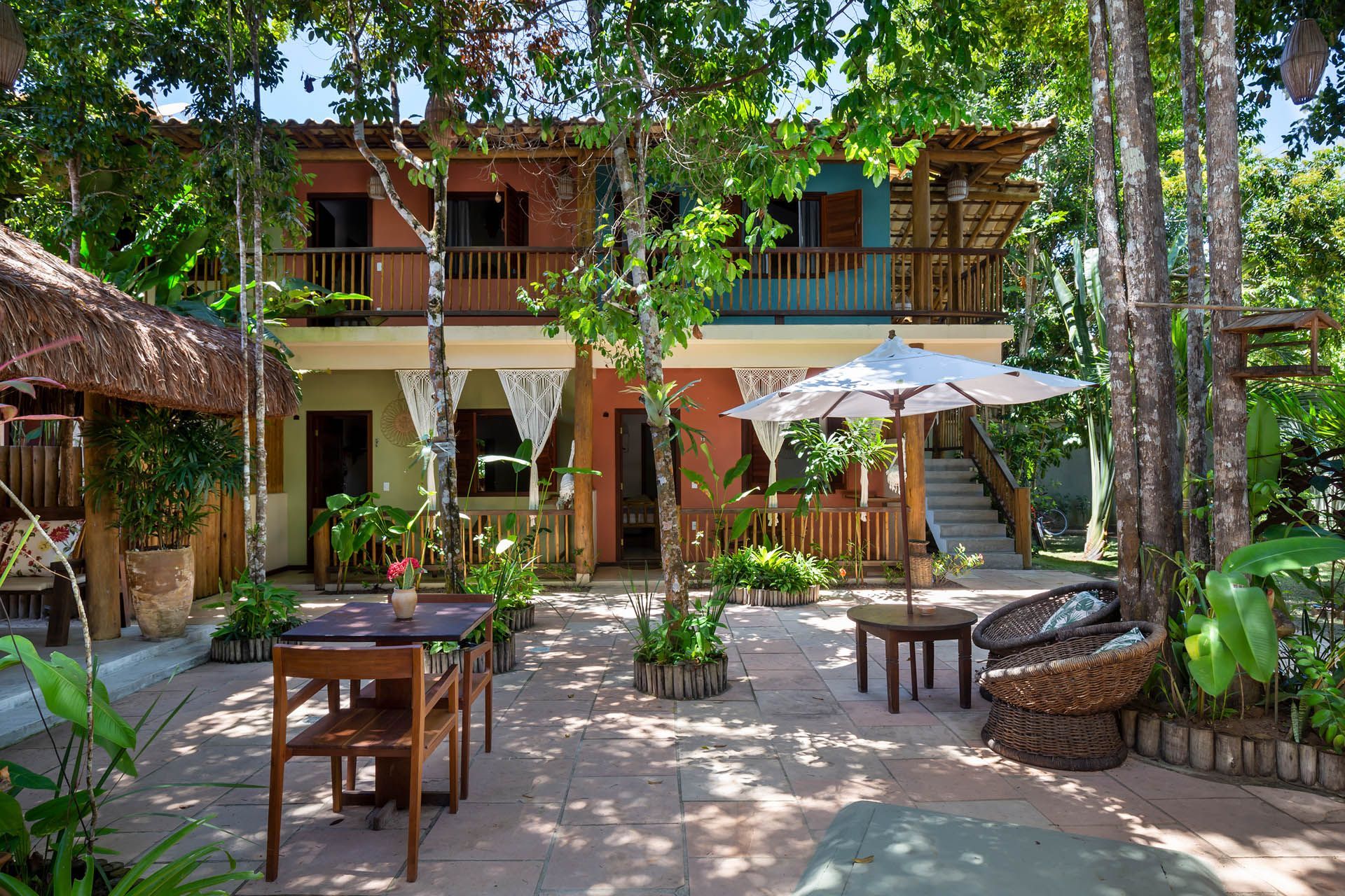 Pátio tropical com uma casa de madeira de dois andares, mesas, cadeiras, guarda-sóis e vegetação exuberante.