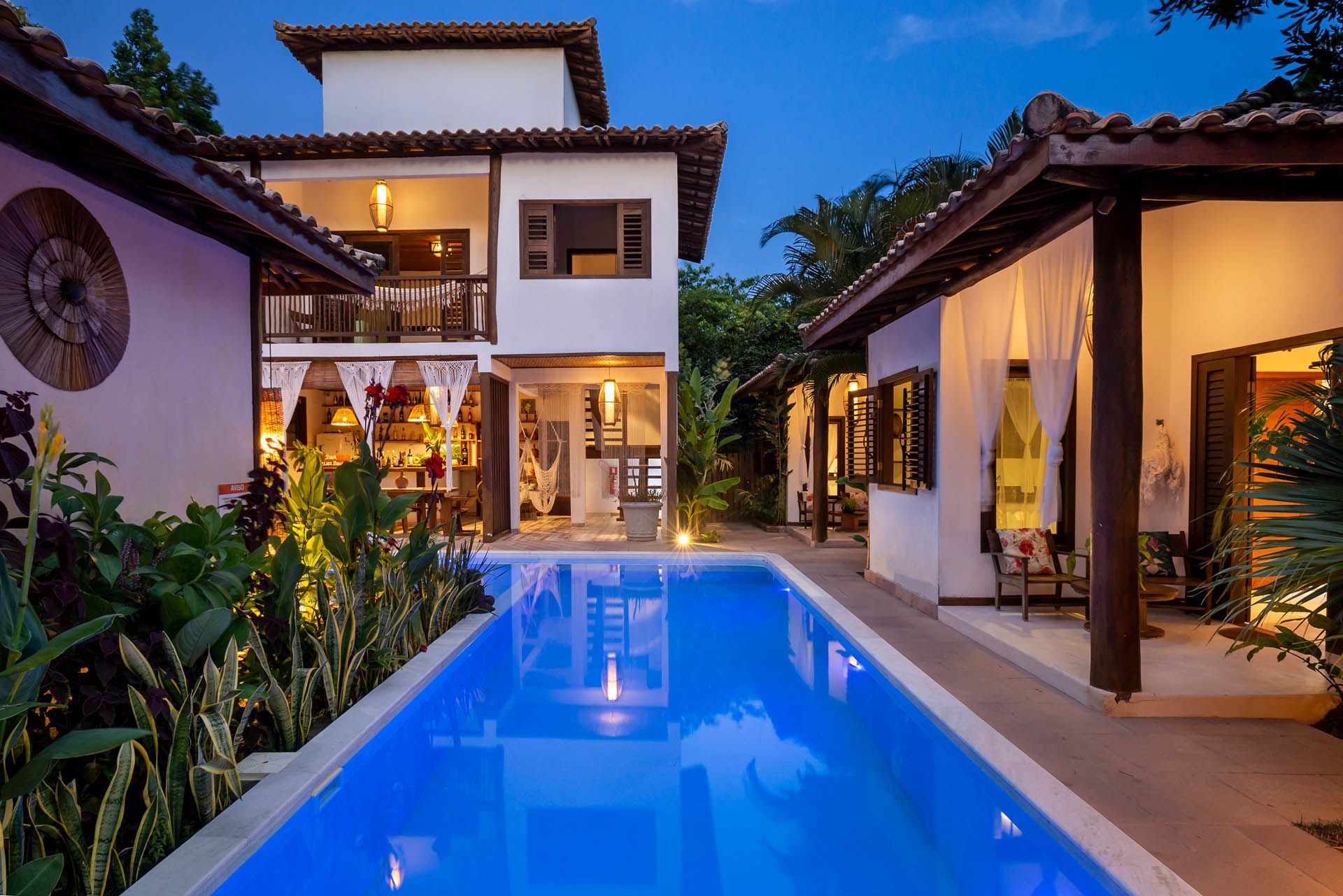 Pátio de uma villa de luxo com uma piscina azul brilhante ao entardecer, emoldurada por varandas com iluminação aconchegante e plantas tropicais.