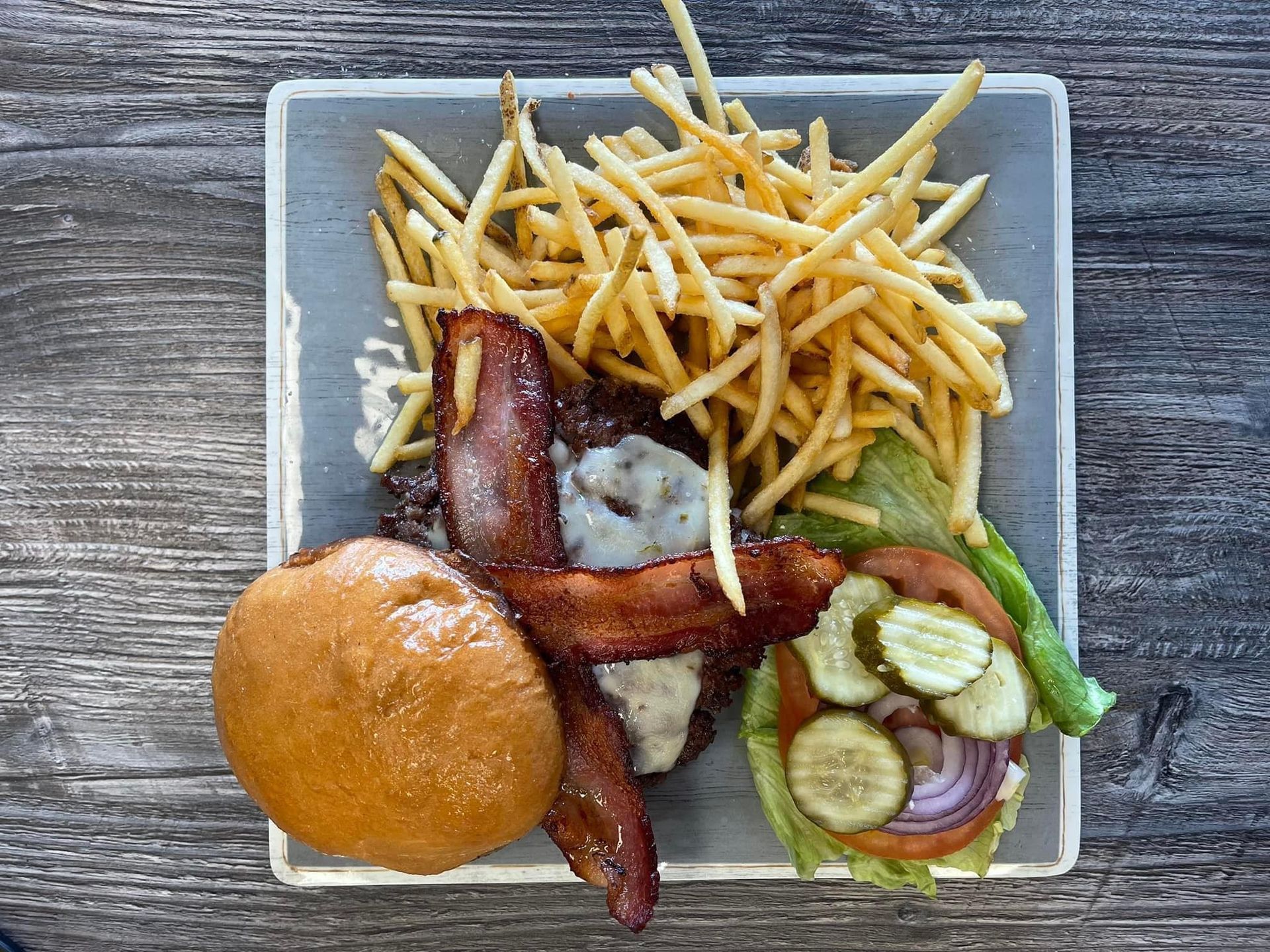Burger, fries, bacon strips, and salad with cucumbers on a square plate.