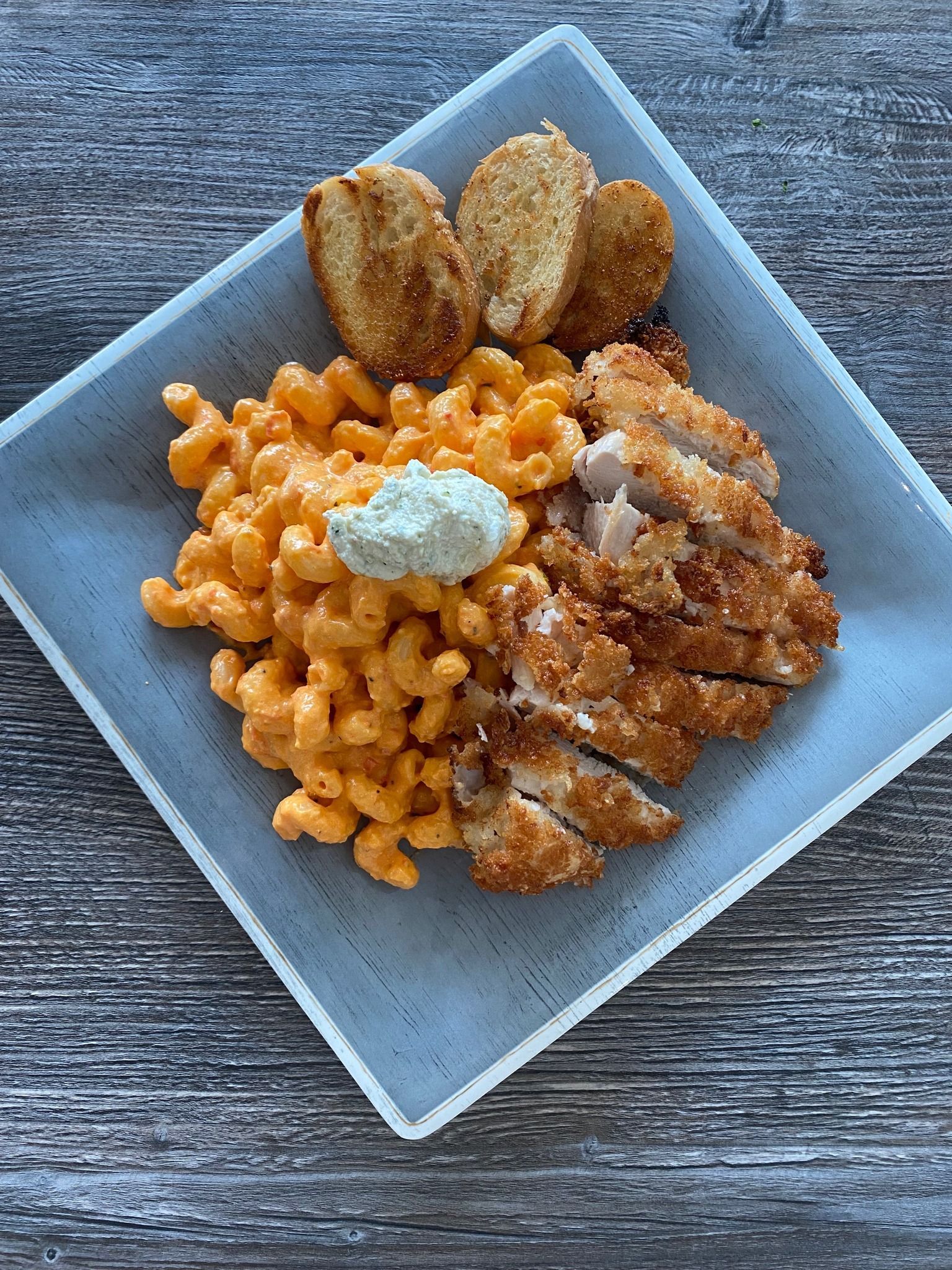 Blue bowl of cheesy pasta with sliced chicken and breaded pieces on a wooden table.