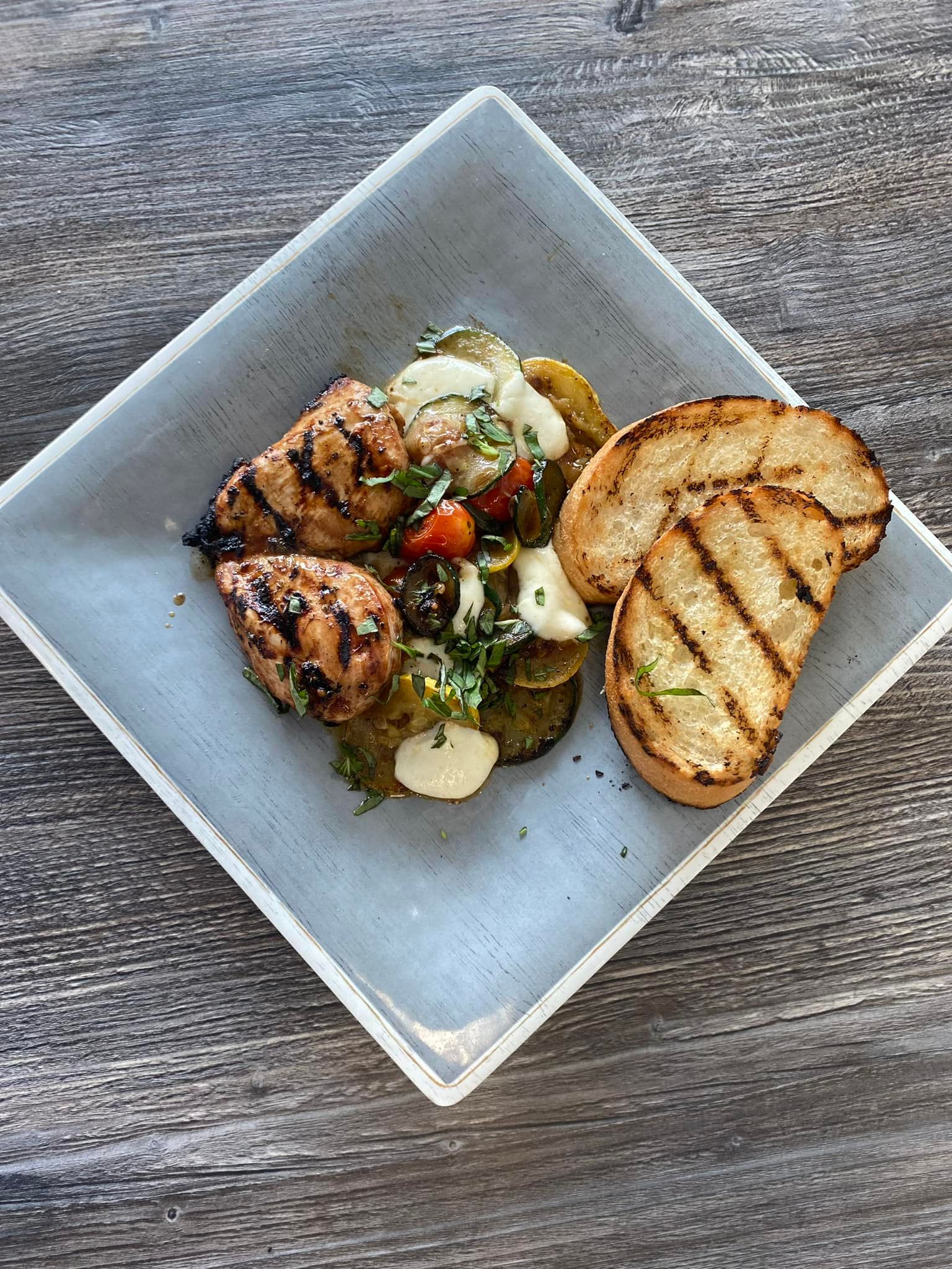 Plated grilled chicken and vegetables with toasted bread on a square white dish.