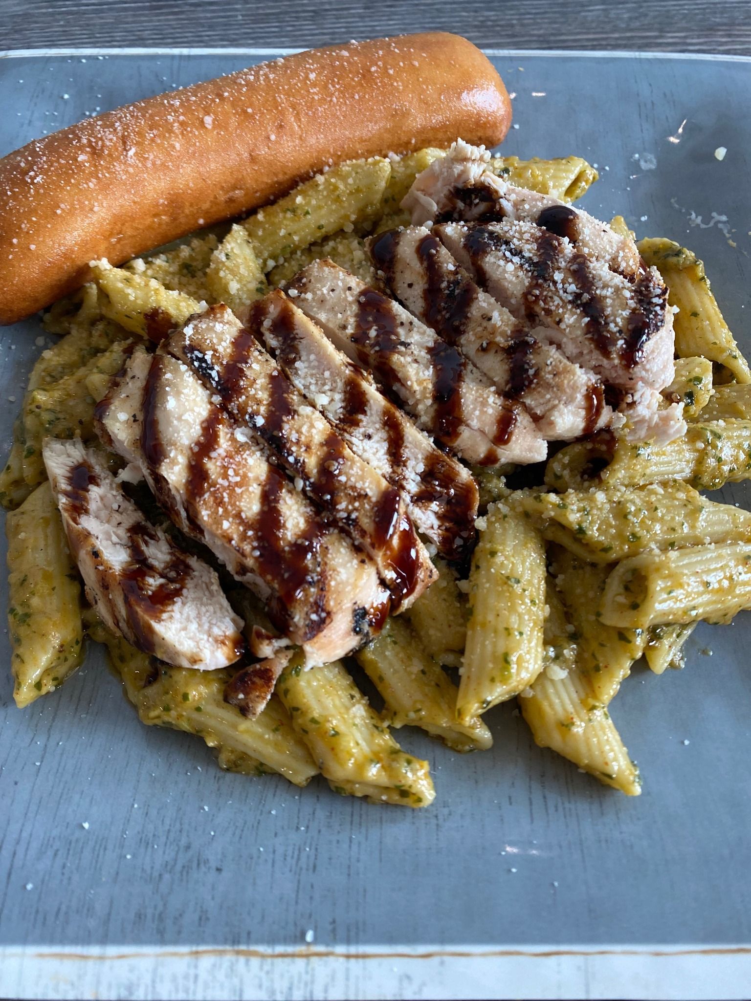 Grilled chicken over pesto pasta with a breadstick on a blue plate