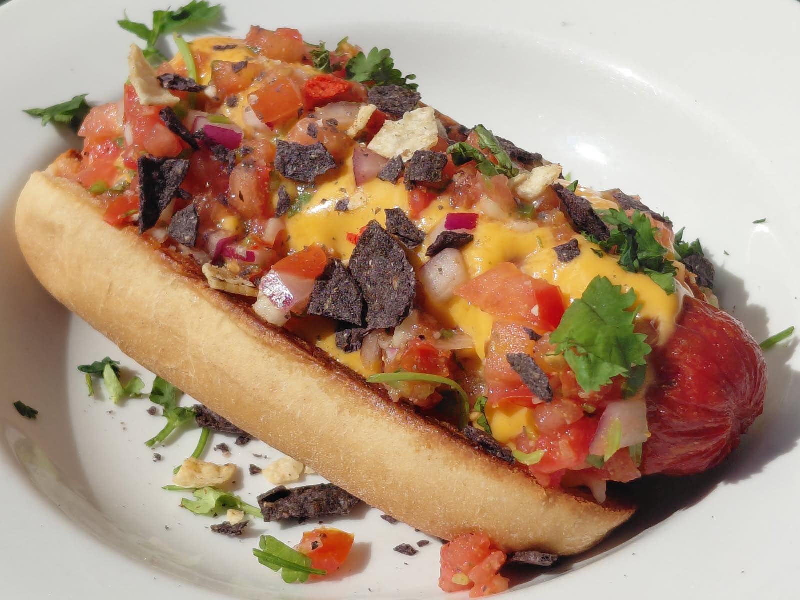 Hot dog topped with colorful diced vegetables, sauce, and herbs on a white plate
