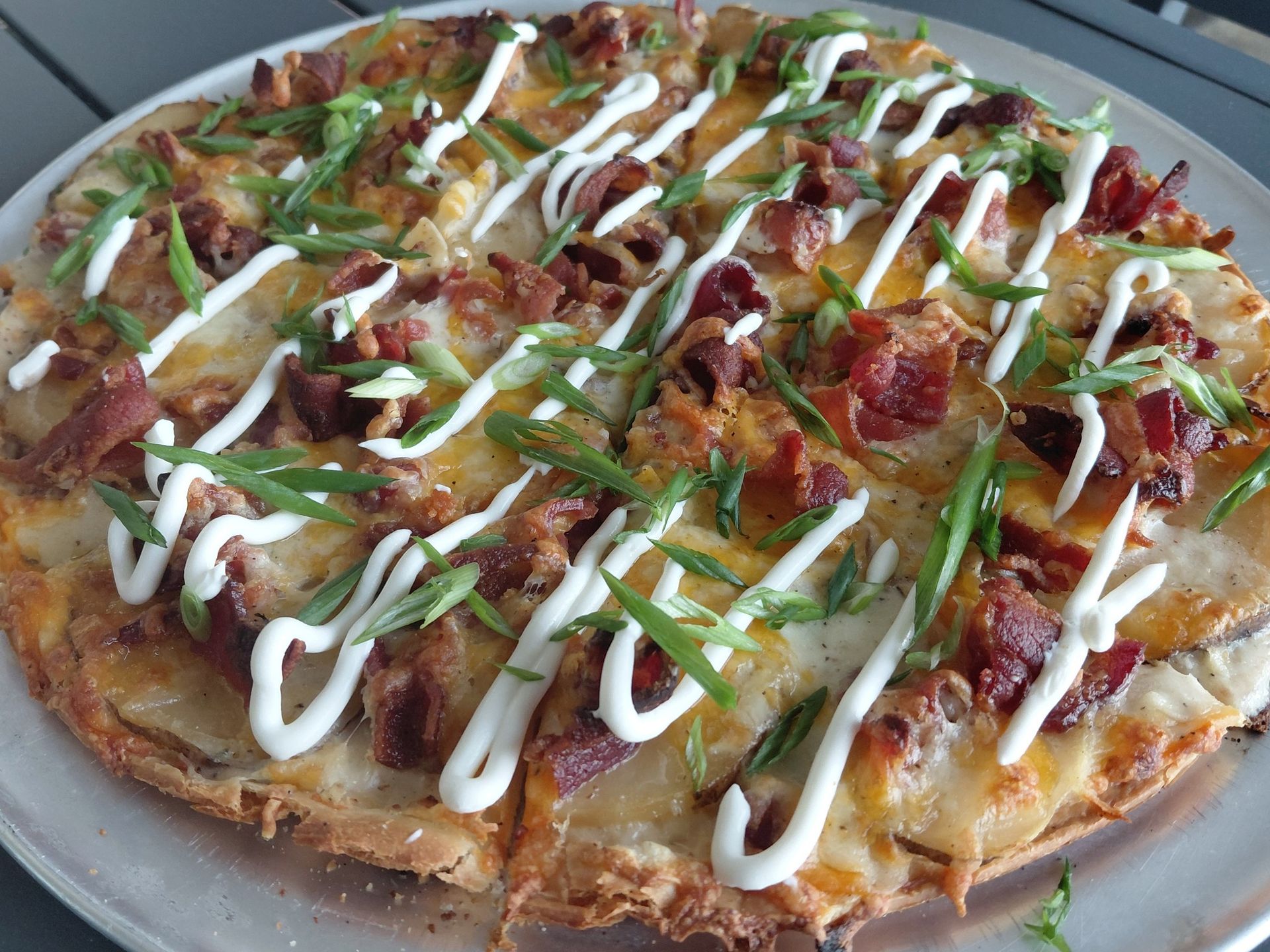 Topped pizza drizzled with white sauce, green onions, and bacon on a plate