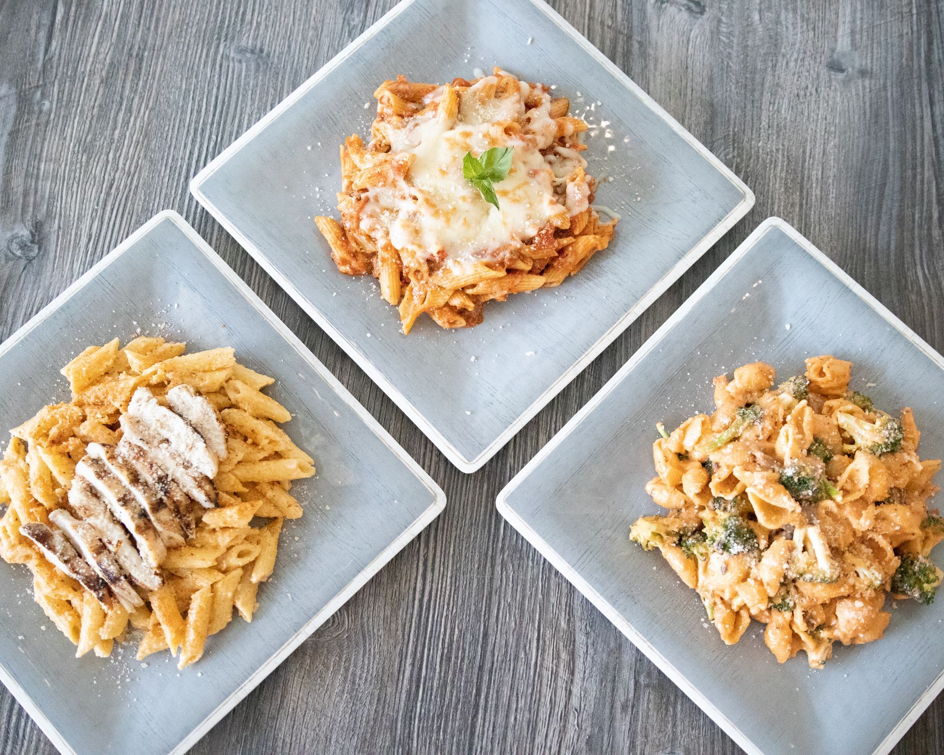 Three square plates of pasta dishes on a wooden table, each with different sauce and garnish.