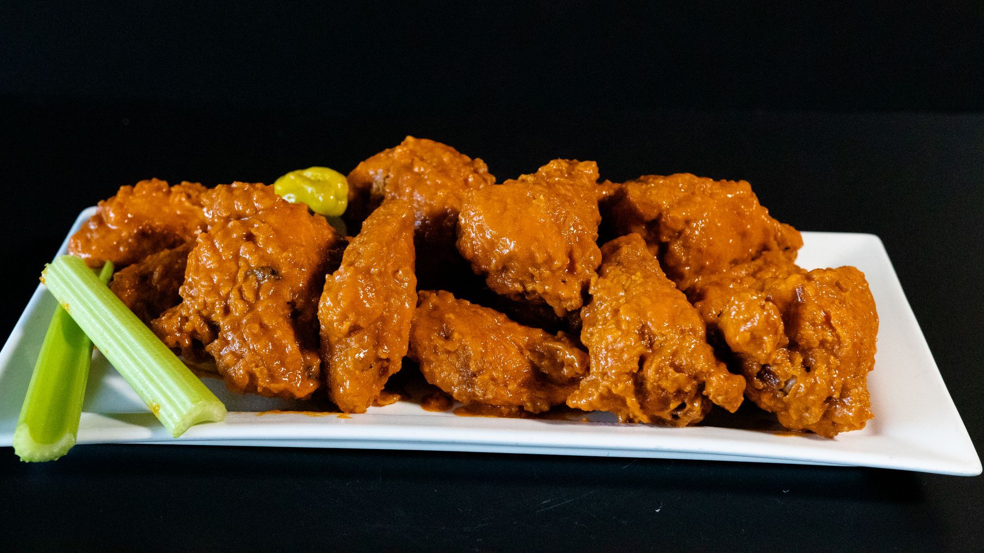 Breaded chicken wings on a white plate with celery sticks and a lime wedge on a black background