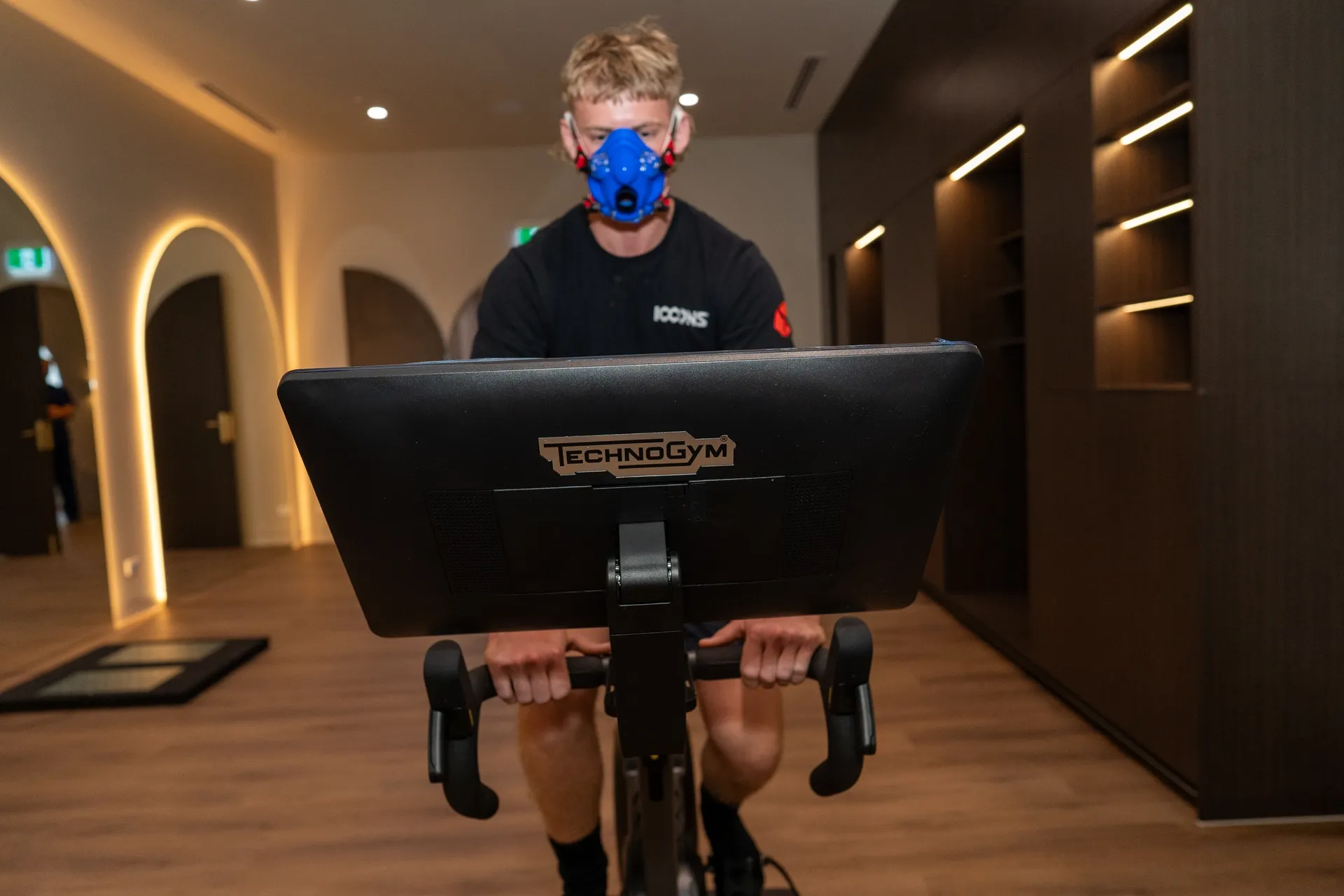 Person wearing a mask cycles indoors on a stationary bike, facing a monitor.