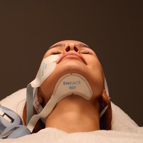 Woman undergoing Emface treatment, lying down with device pads on her face and neck.