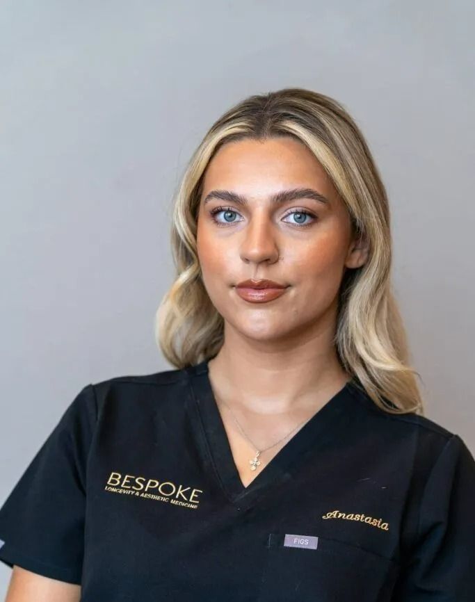 Woman wearing black scrubs with blonde hair, looking directly at the viewer, small gold necklace.