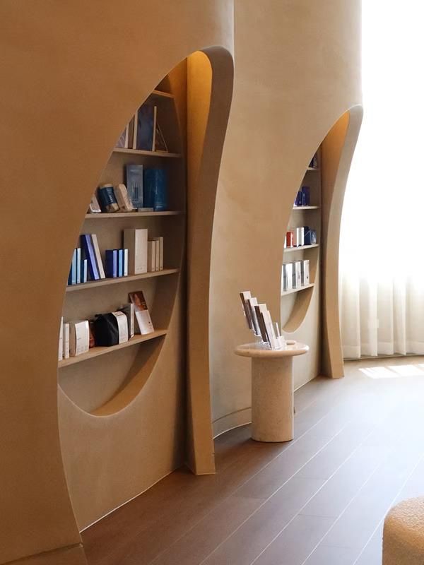 Beige bookshelves recessed into a curved wall. Books and displays on shelves. Wooden floor, soft lighting.
