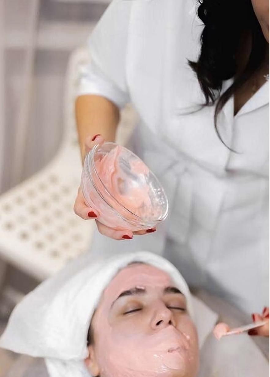 Person receiving facial treatment with pink mask. Therapist holding mask product.