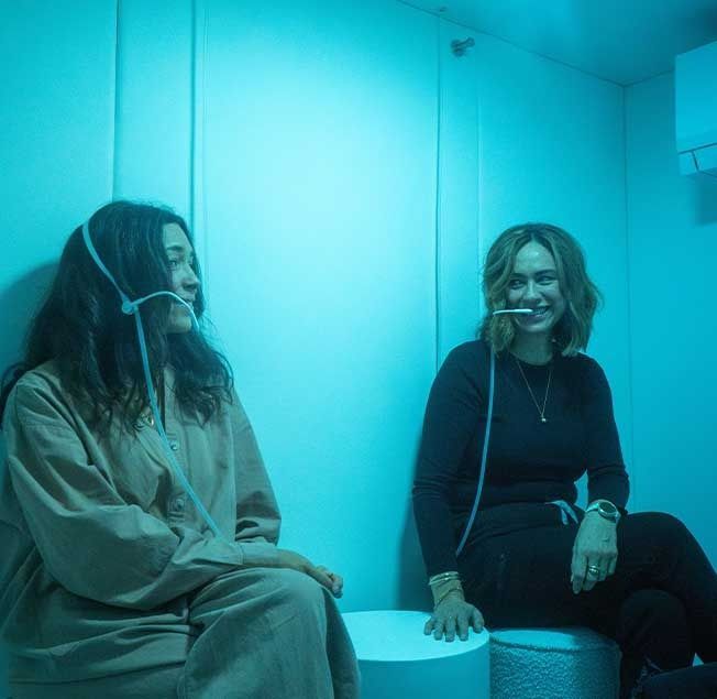 Two people inside a blue-lit room, each with a tube in their mouths. One smiles.