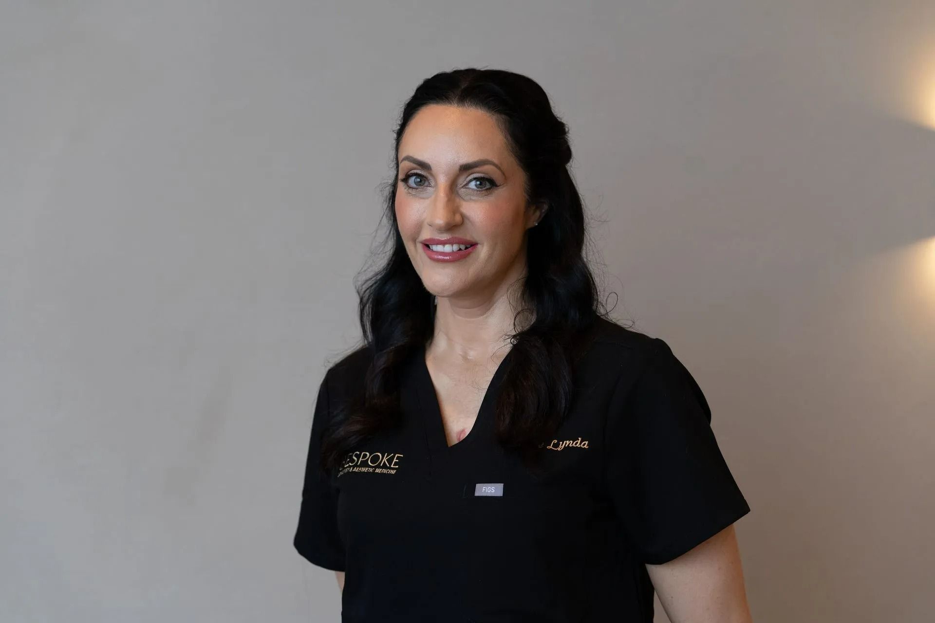 Woman in black scrubs smiles at the camera, standing in front of a neutral-colored wall.