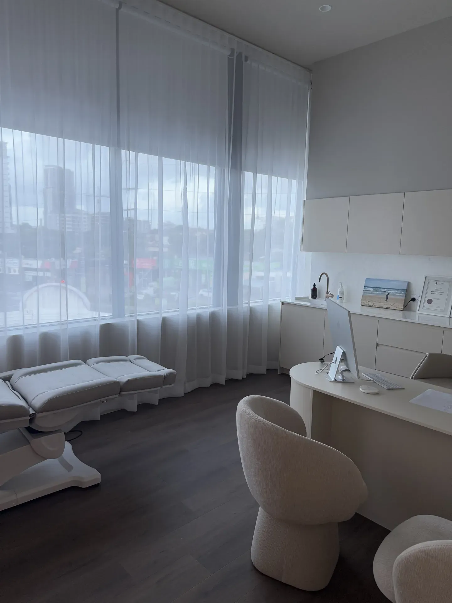 Bright medical office with large windows, exam table, desk, and two chairs.