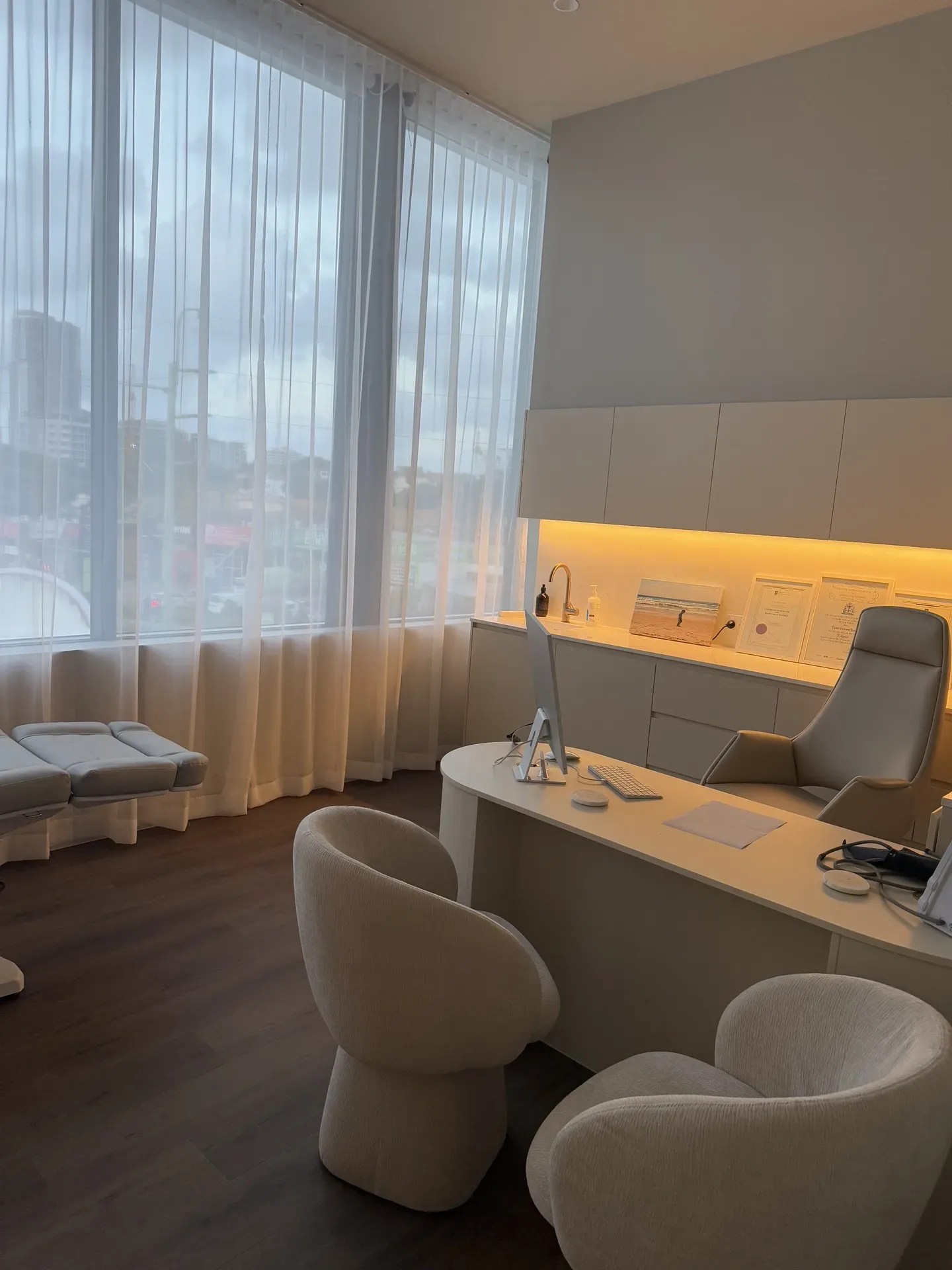 Modern medical office interior with curved desk, cream chairs, treatment bed, and large window.