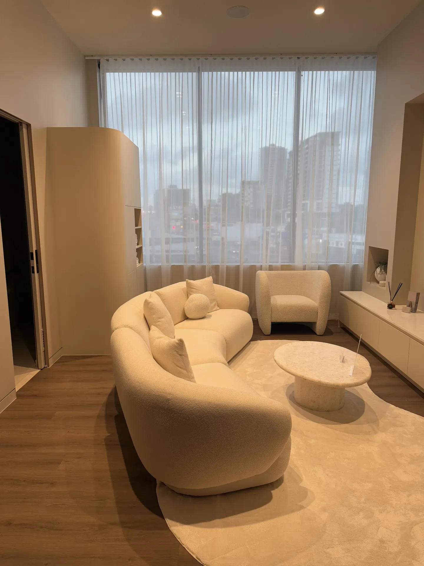 Cream-colored living room with curved sofa, armchair, coffee table, sheer curtains, and light wood floors.